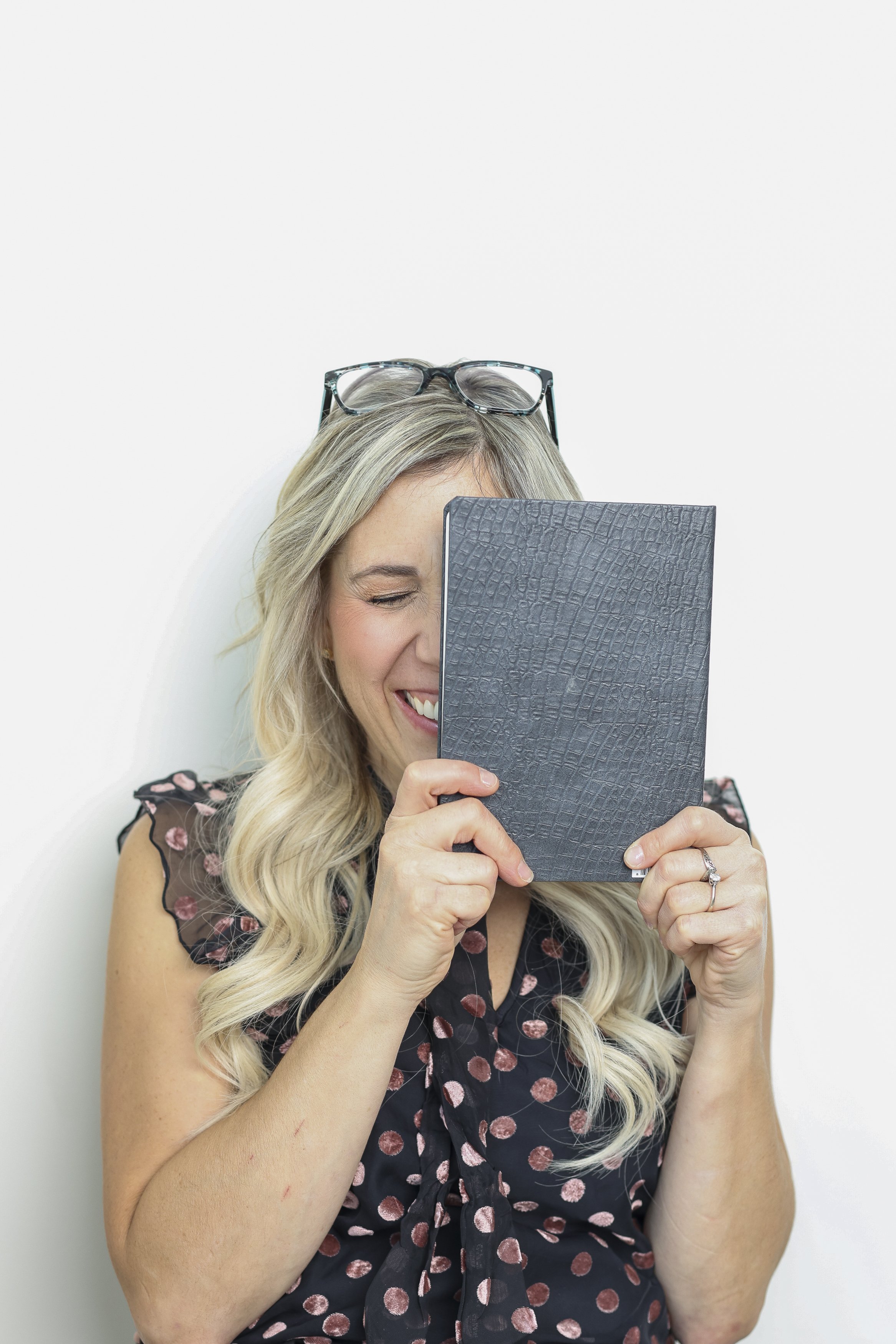 A woman with long blonde hair, laughing or smiling, holding a black folder or notebook in front of her face. She is wearing glasses on her head and a black dress with pink polka dots.