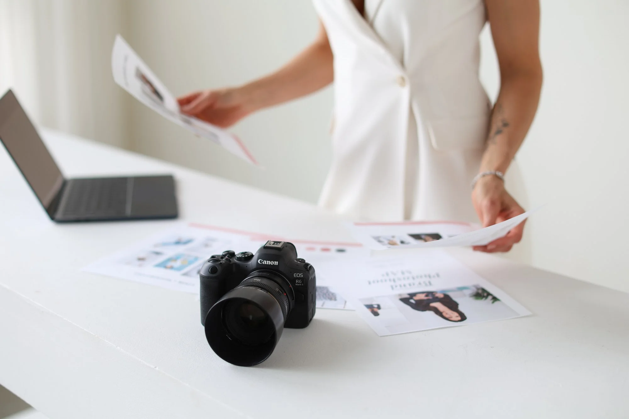 woman holding papers with laptop on table and camera in sight, wearing white suit