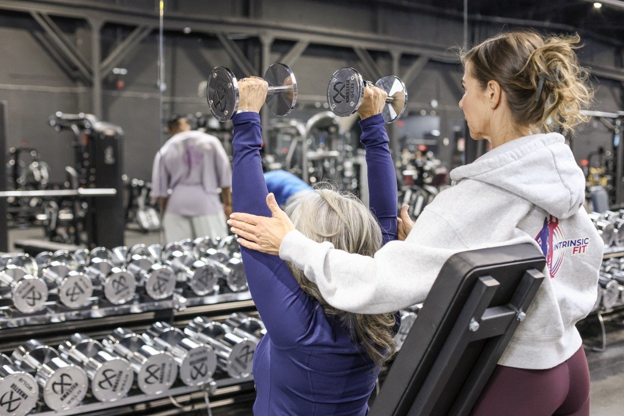 woman spotting another woman lifting dumbells overhead in a gym