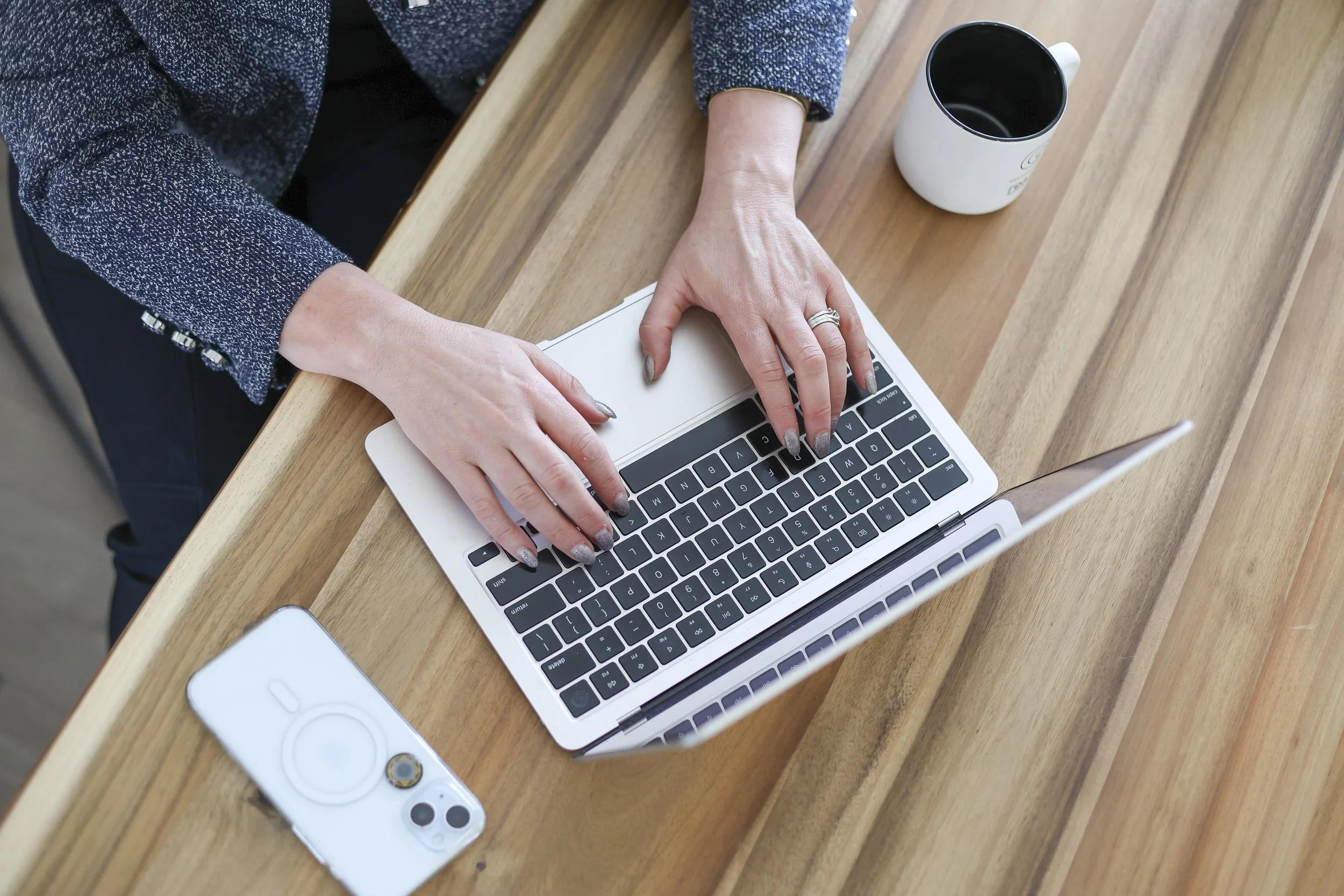 Person using a laptop on a wooden desk, with a cup and a phone nearby.