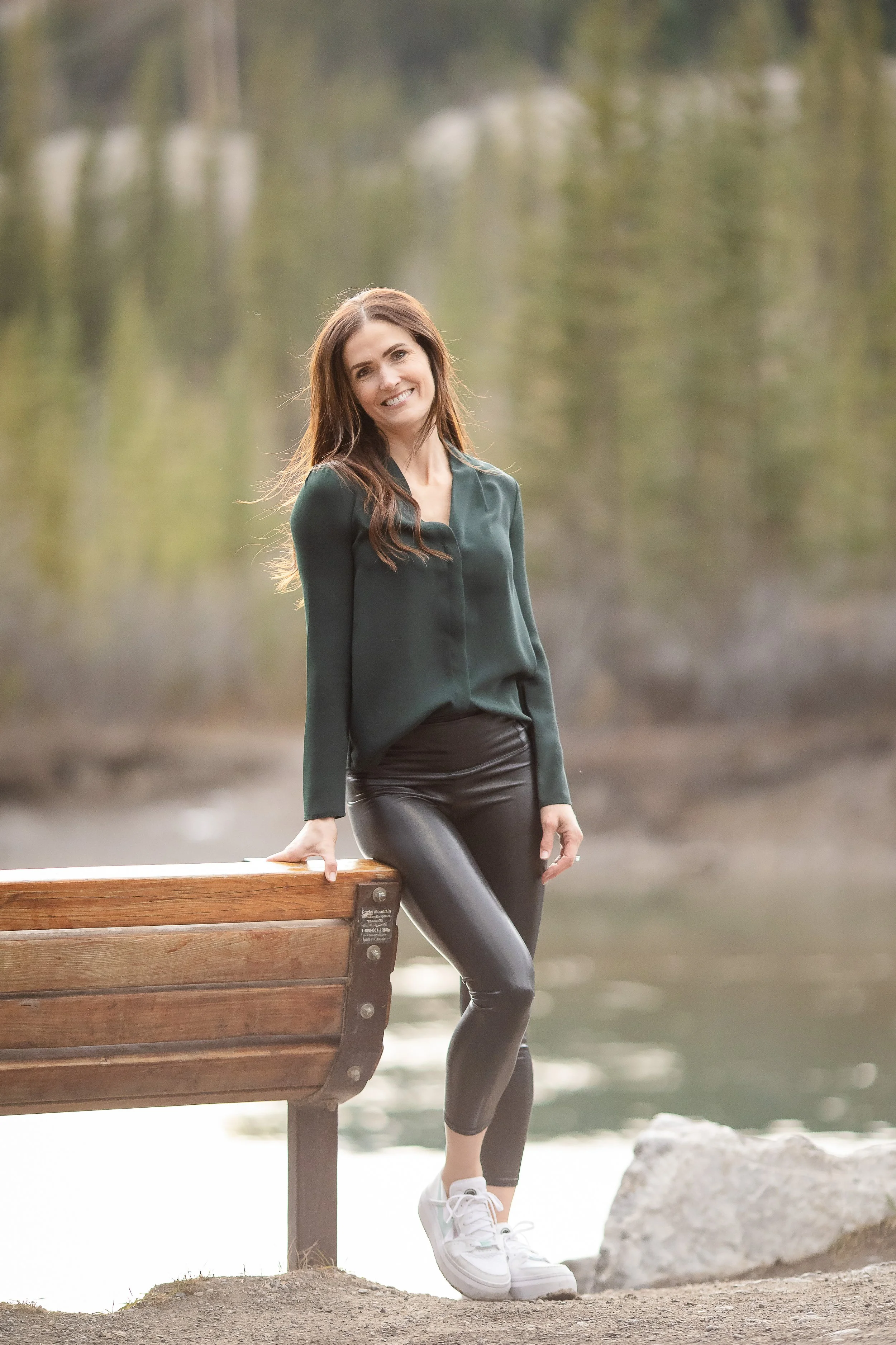 A woman with long brown hair smiling, wearing a dark green blouse, black shiny leggings, and white sneakers, standing by a wooden bench near a body of water with a forested background.