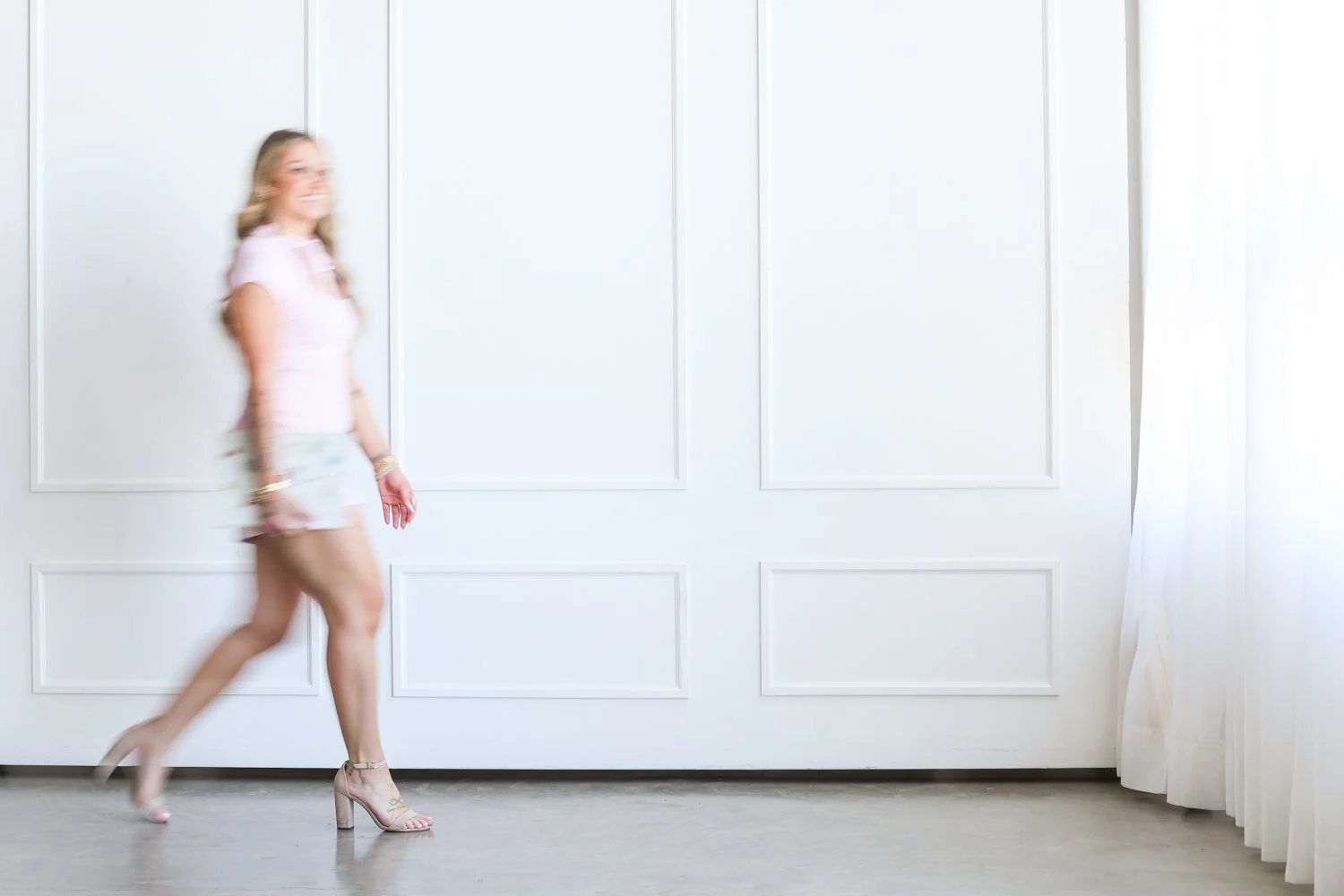 wman in a pink dress holding a laptop walking toward a window blurred to show movement against a white wall