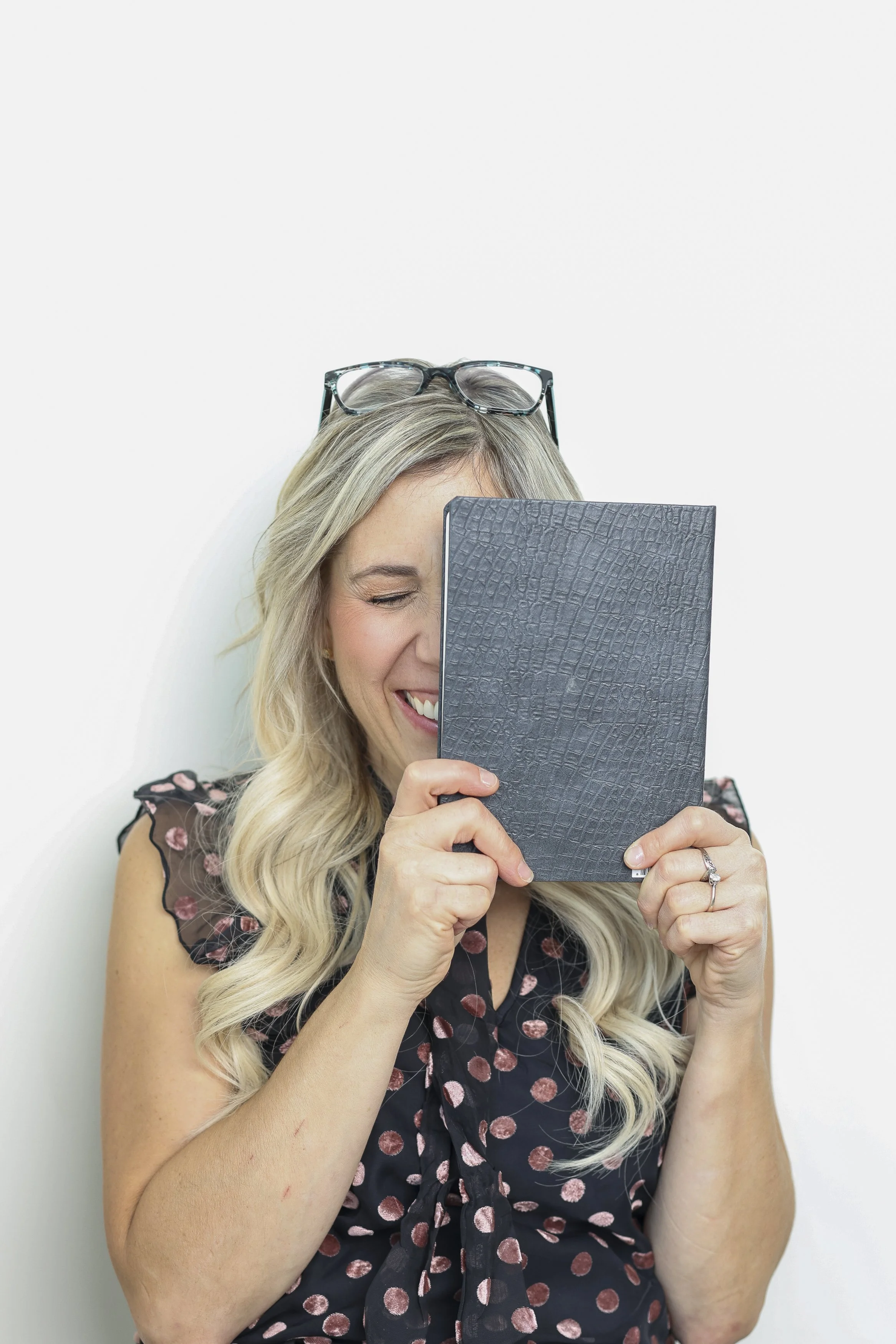 Woman holding a black textured notebook, smiling and laughing with glasses on her head, wearing a polka dot blouse, against a plain white background.