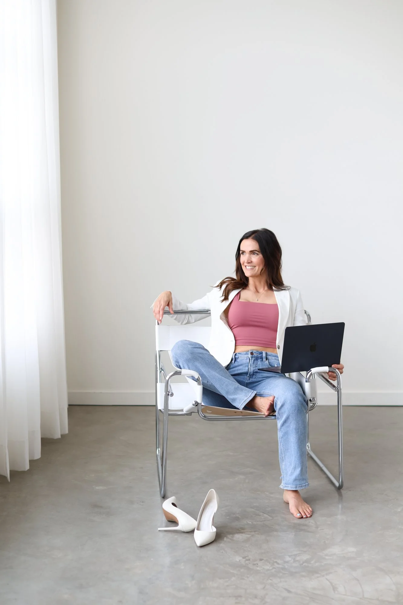 woman sitting casually on chair with laptop in lap looking out the window, one leg up on the chari and heels casually left on the floor, smile on her face in her pink top with white blazer and jeans