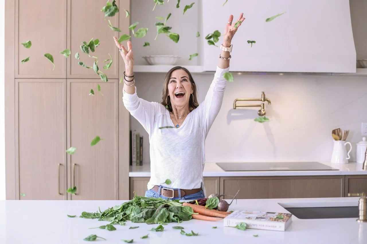 woman throwing spinach in the air with a smile on her face in the kichen withvarious vegetables on the counter