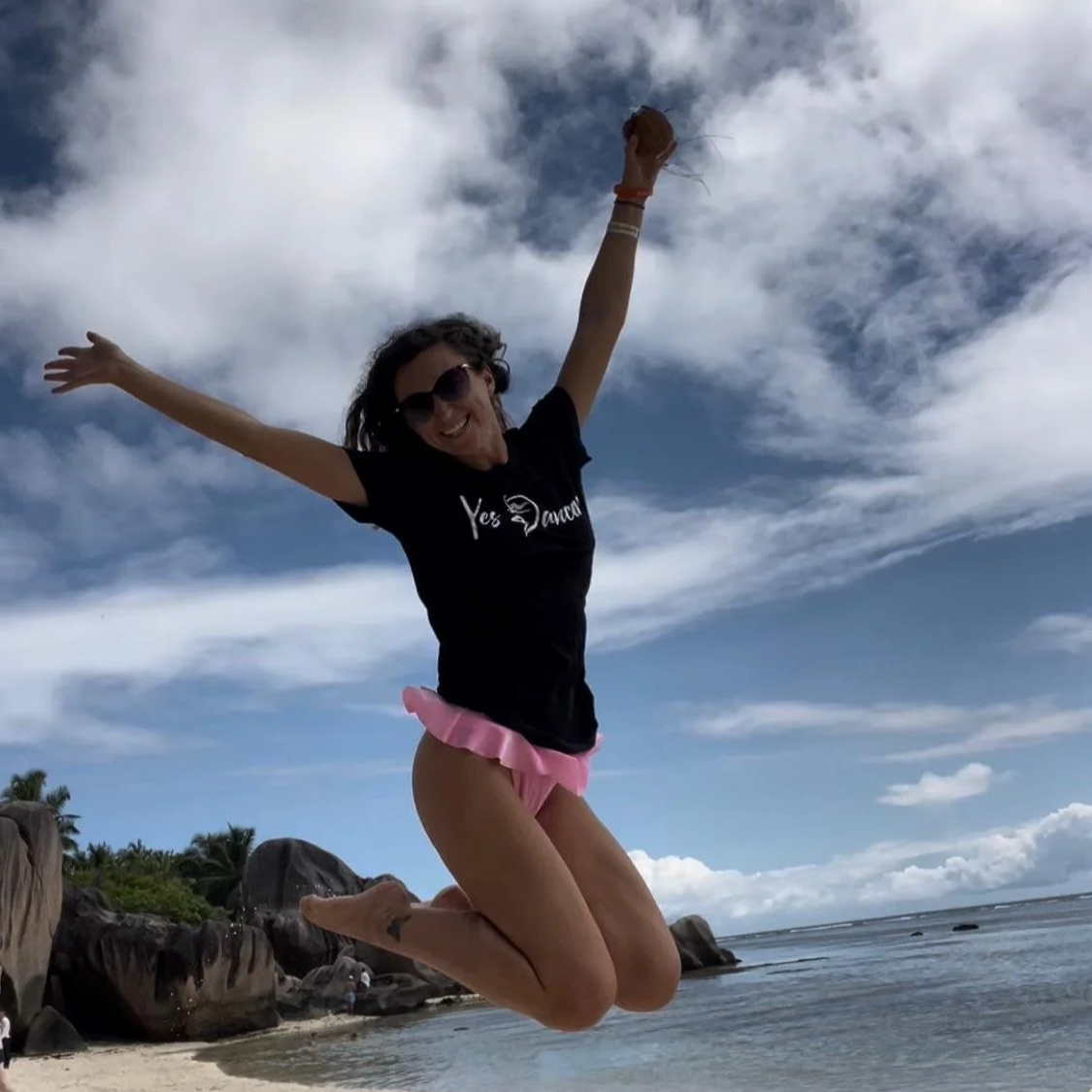 A woman jumping joyfully on a beach with rocks and palm trees in the background, wearing sunglasses, a black t-shirt, and pink shorts, under a partly cloudy sky.