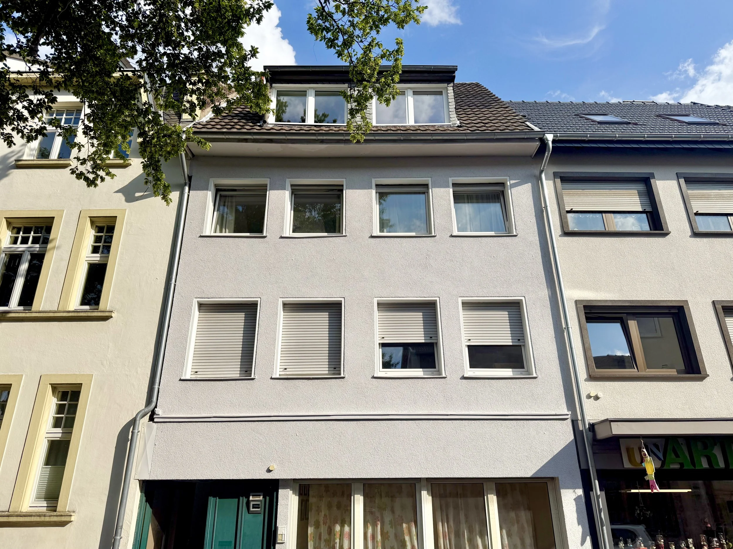 Mehrstöckiges Wohnhaus mit weißen und dunklen Fensterrahmen, Dach mit Dachfenstern, Baum und blauer Himmel im Hintergrund.