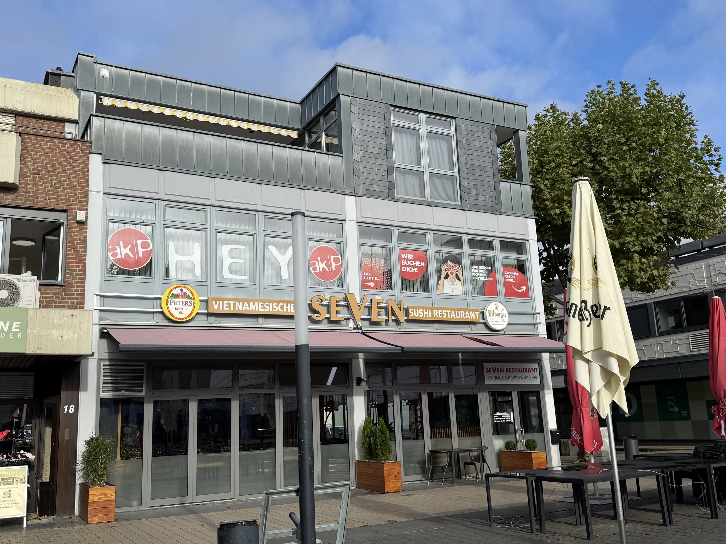 Ein zweistöckiges Gebäude mit einem vietnamesischen Sushi-Restaurant namens SEVEN, große Fenster mit Dekorationen, Außenbestuhlung mit Tischen und Stühlen, Sonnenschirme, Bäume und ein blauer Himmel.