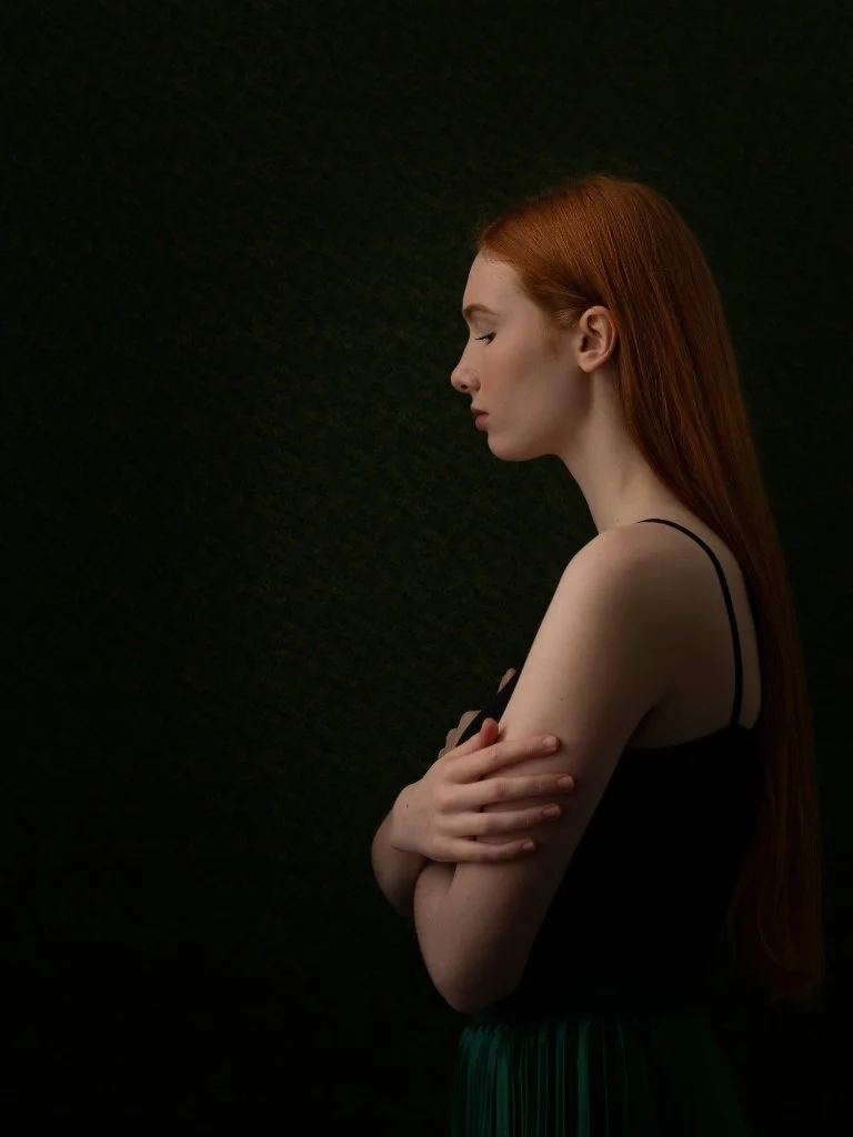 Side profile of a woman with long red hair, wearing a black sleeveless top, standing against a dark background with her arms crossed