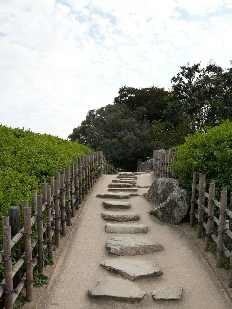 Stone pathway with uneven flat stones, bordered by wooden fences and green bushes, leading towards trees in the distance under a partly cloudy sky.