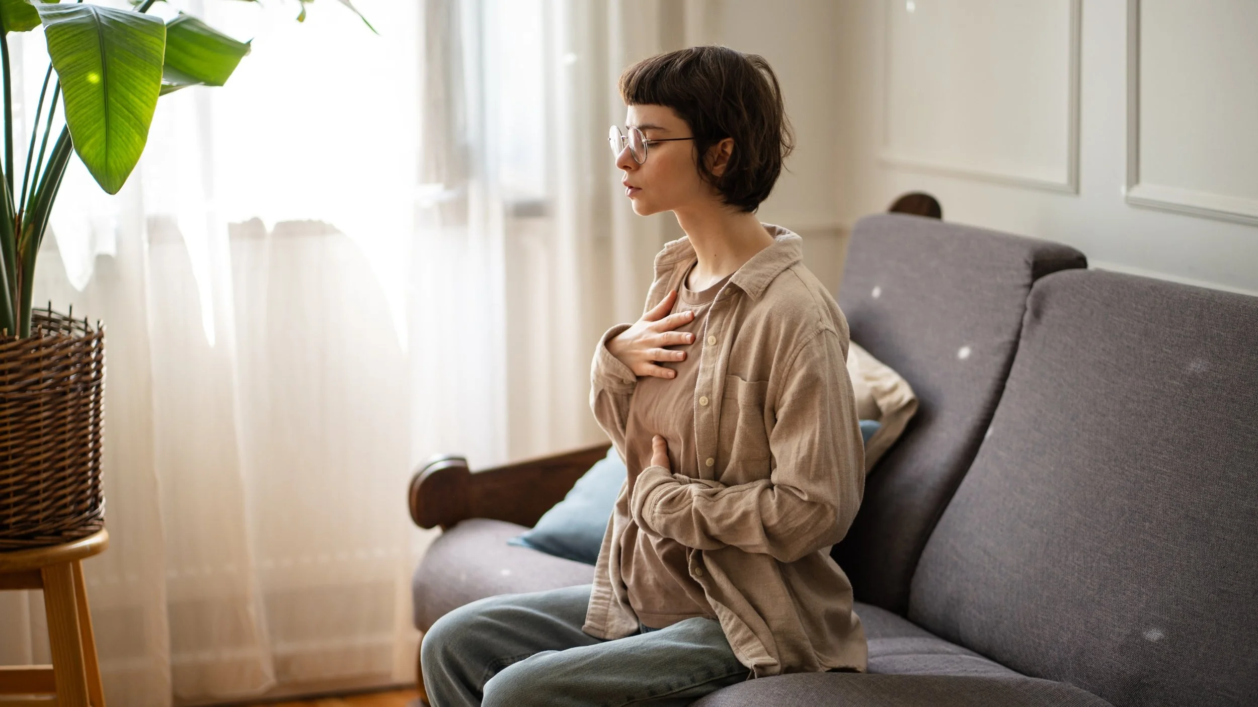 A young woman with short dark hair, glasses, and wearing a beige button-up shirt sitting on a gray sofa in a well-lit room, holding her hand on her chest and stomach with a concerned expression.