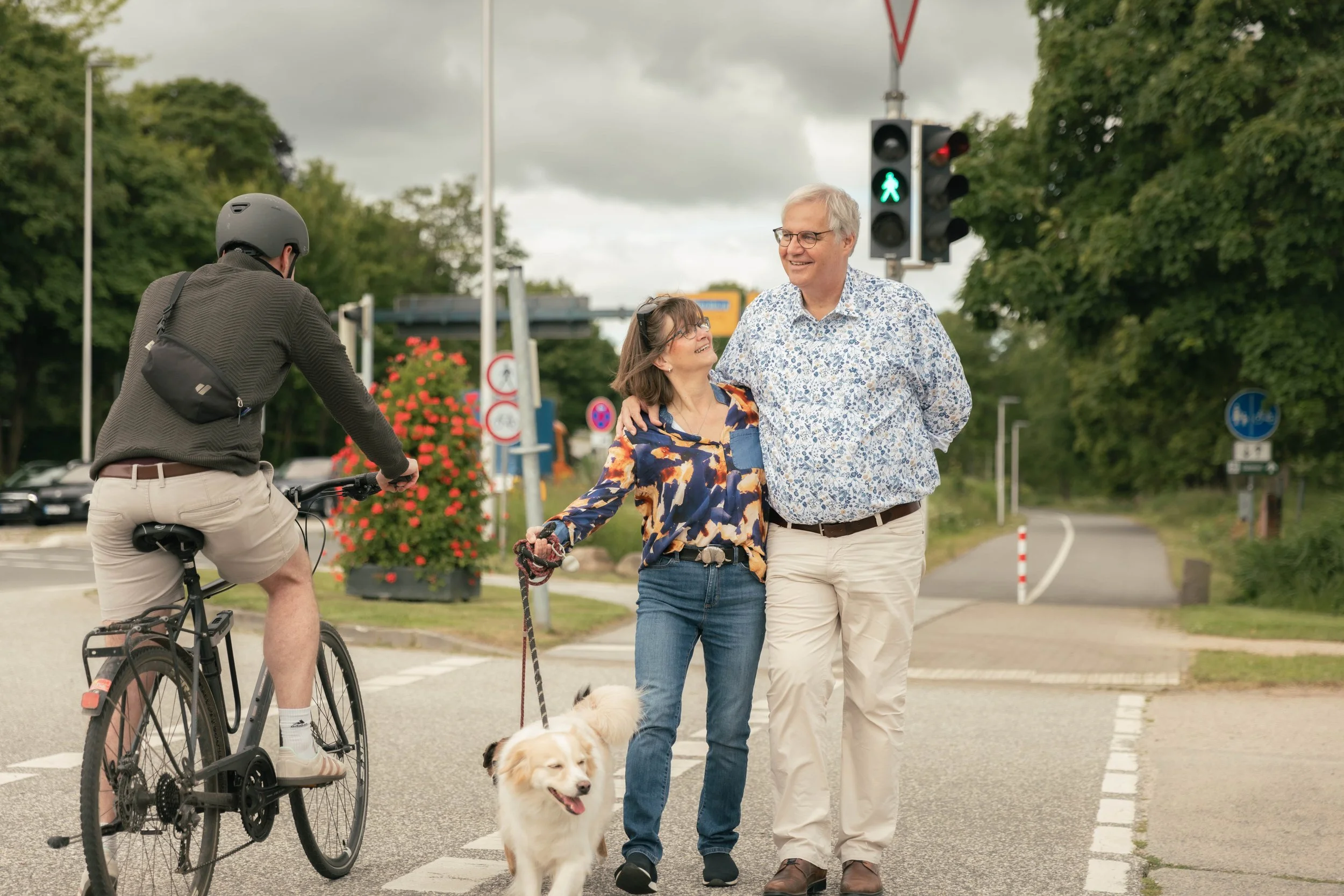 Ein älteres Paar geht Hand in Hand mit einem Hund über eine Fußgängerampel, während ein Radfahrer vorbeifährt. Das Paar lächelt und scheint sich zu freuen. Es ist ein bewölkter Tag und im Hintergrund gibt es grüne Bäume und Verkehrsschilder.