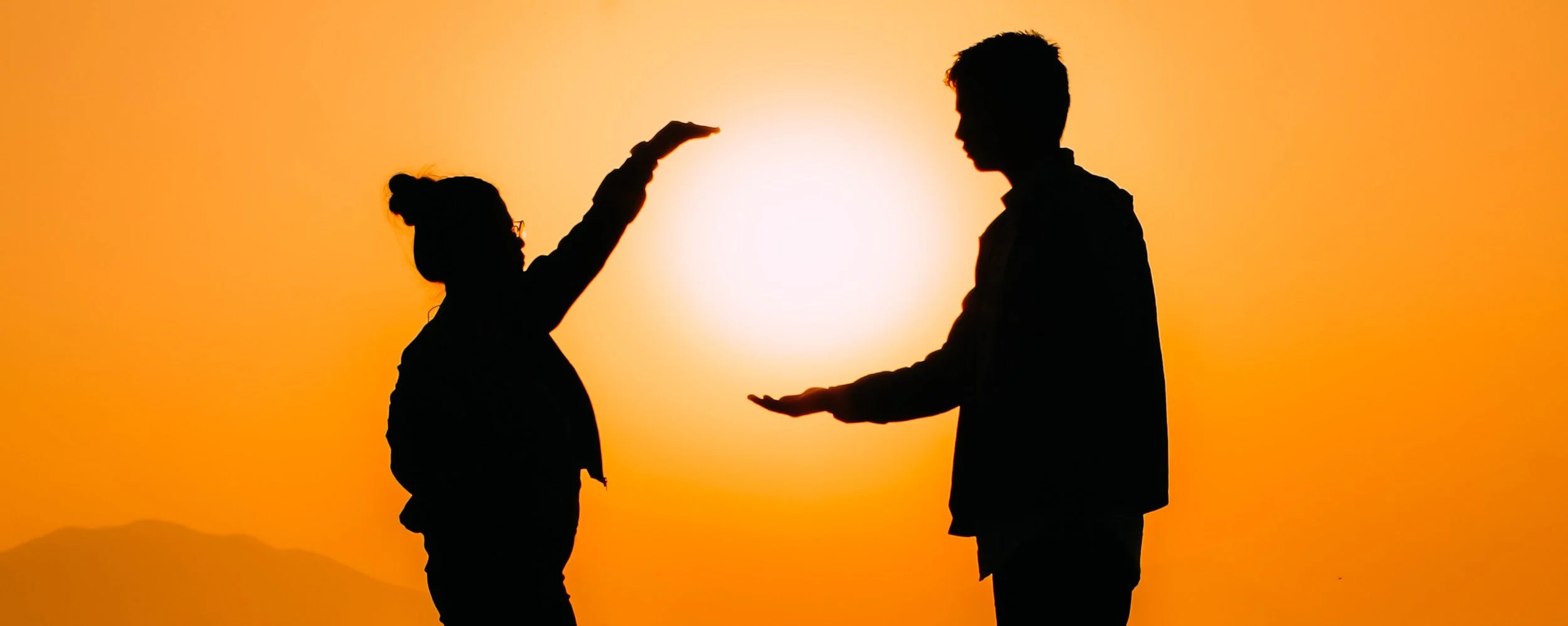 Silhouettes of a woman and a man against an orange sunset, with the woman placing her hand above the man's head and the man holding his hand open in front of him.