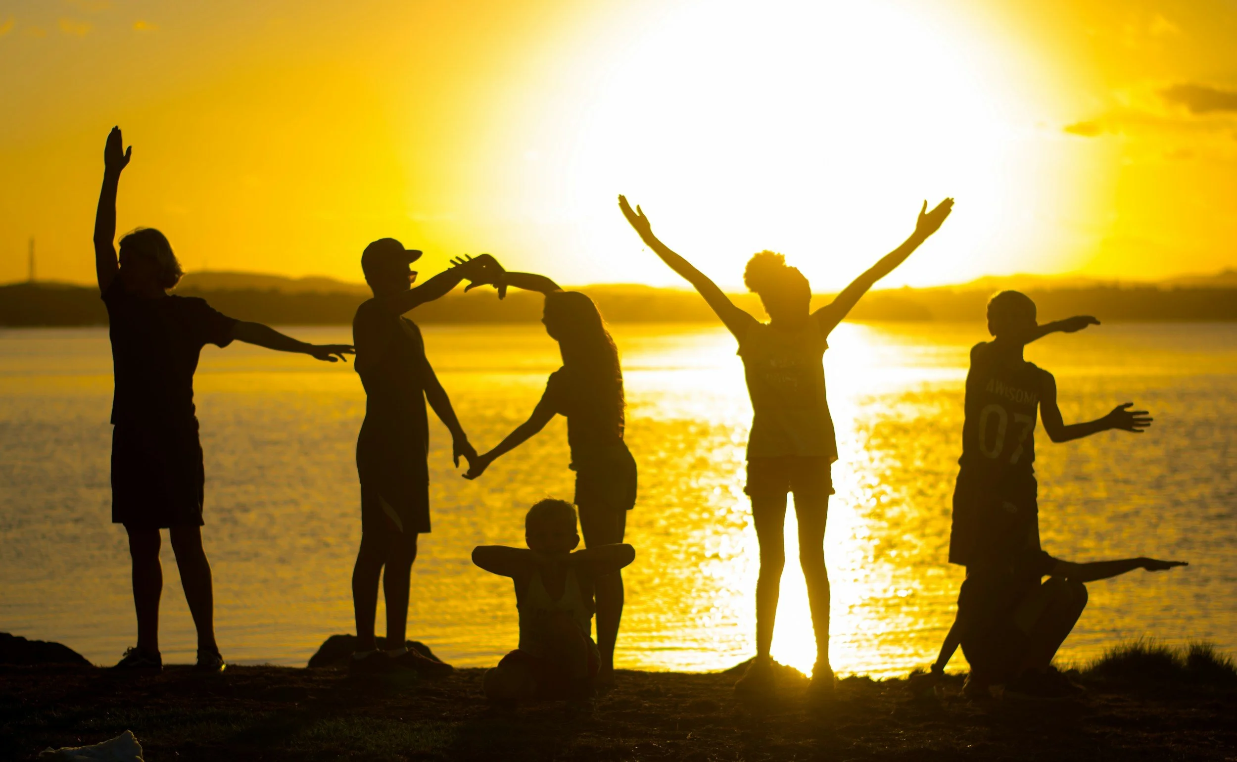 Silhouettes of children playing and holding hands by a body of water at sunset.