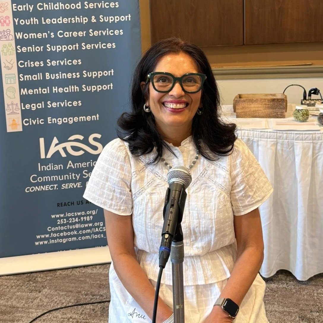 Anu smiles broadly while seated in front of a microphone. She is wearing a white blouse and skirt and there is a sign about the Indian American Community Services program behind her.