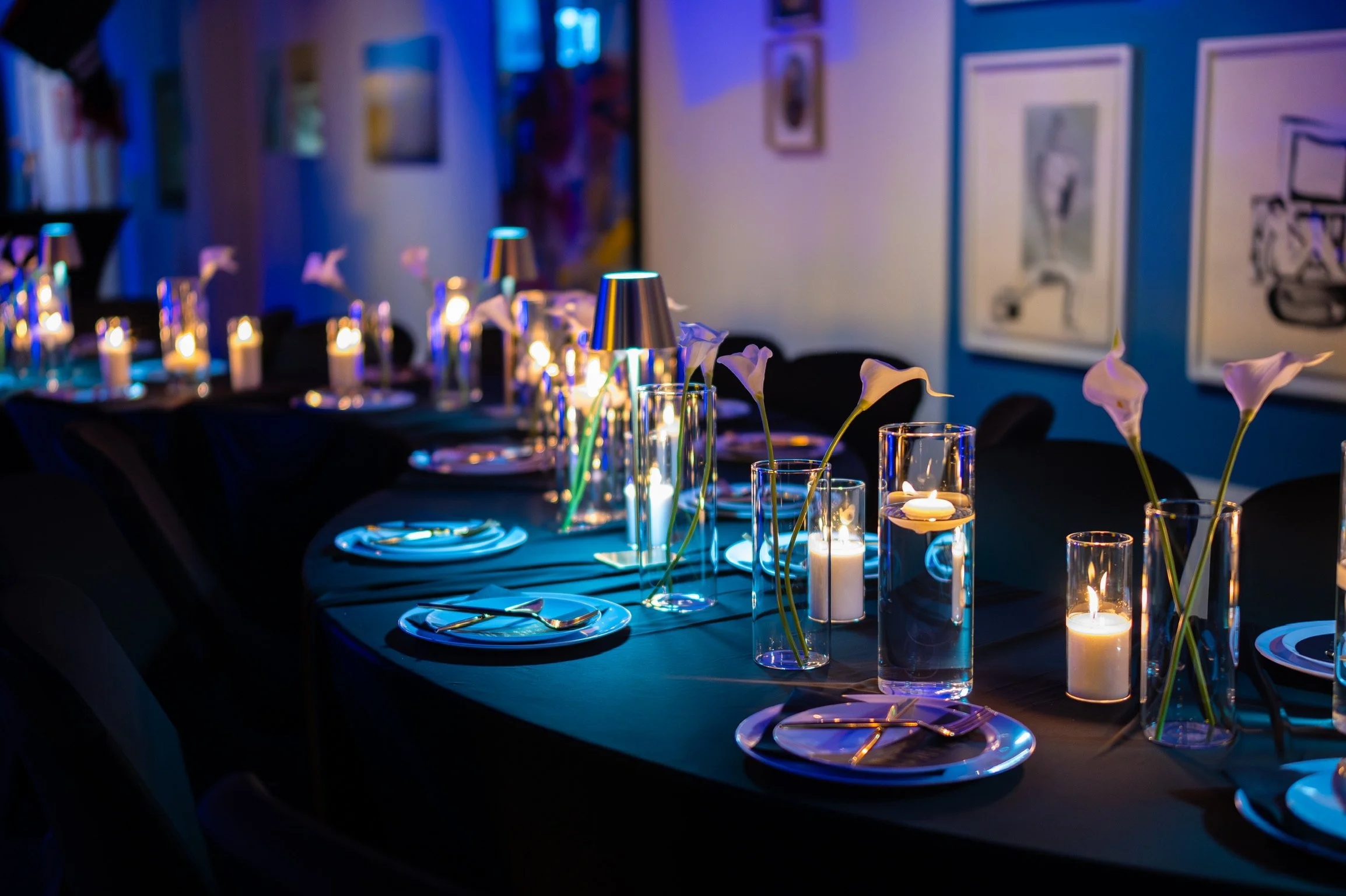 A dimly lit dining table set for a formal dinner with candles, glasses, and flowers, with artwork on the walls in the background.