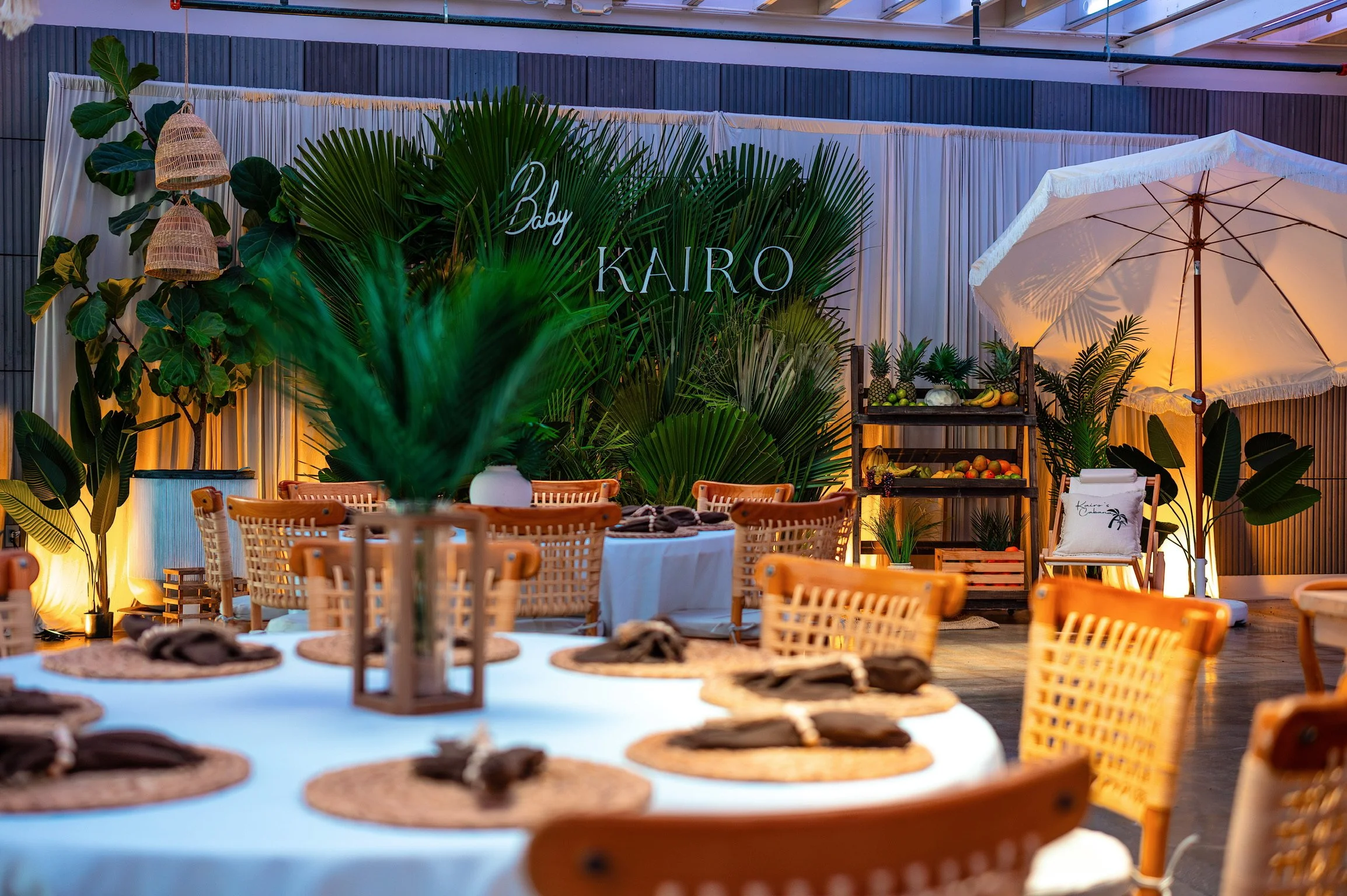 Decorative event setup with lush green tropical plants, a white canopy, a table with woven placemats and desserts, and a sign that says "Baby KAIRO" on a backdrop of white curtains and greenery.
