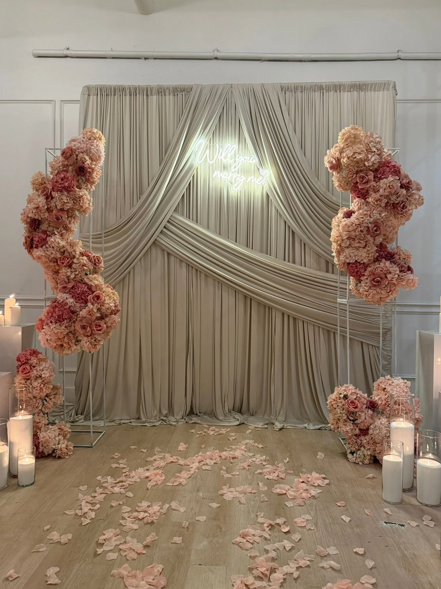 Wedding backdrop with cream-colored curtains, pink flowers, candles, and scattered flower petals on the floor.