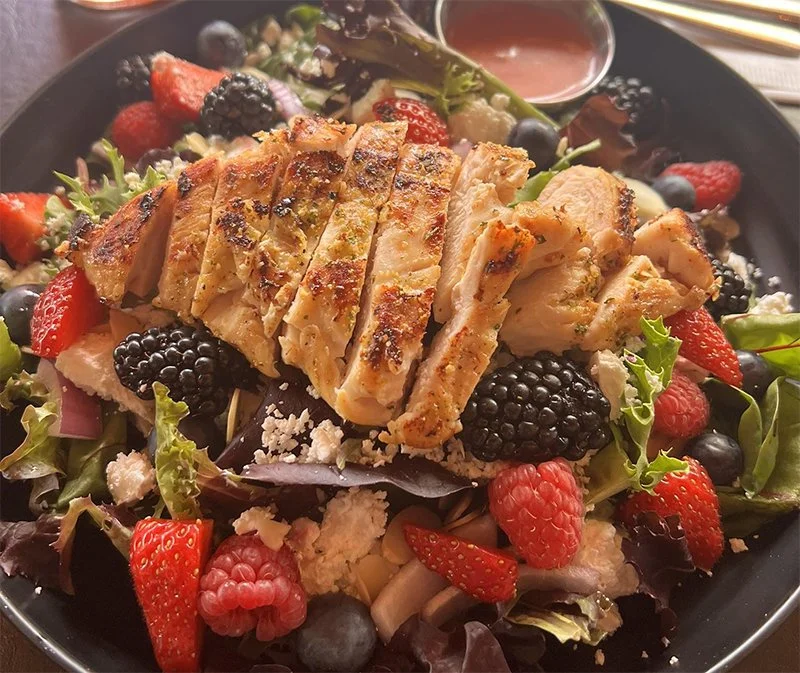Salad with slices of grilled chicken, mixed berries, leafy greens, cheese, and a small container of dressing.