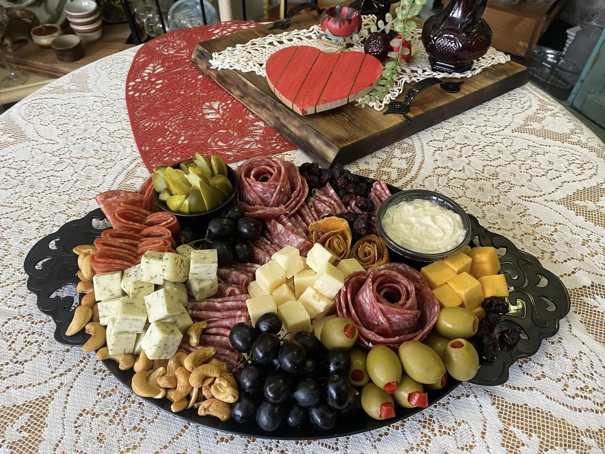 Charcuterie board with cheeses, meats, pickles, olives, nuts, and dried fruits on a table with lace tablecloth, with additional decorative items in the background.