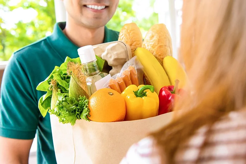 A person hands a grocery bag filled with various fresh fruits and vegetables to another person, with a smiling man in the background.