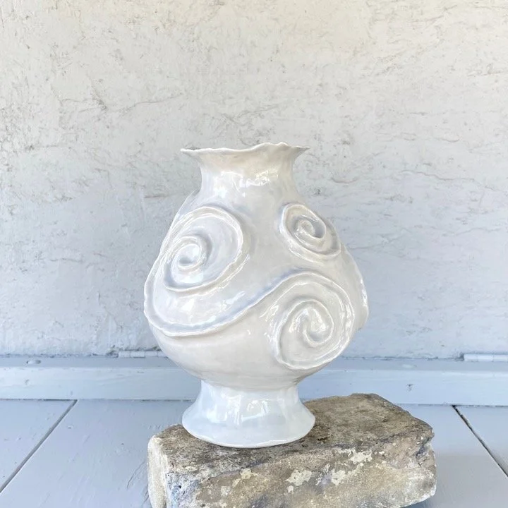 White ceramic vase with spiral designs on a brick, against a white textured wall.