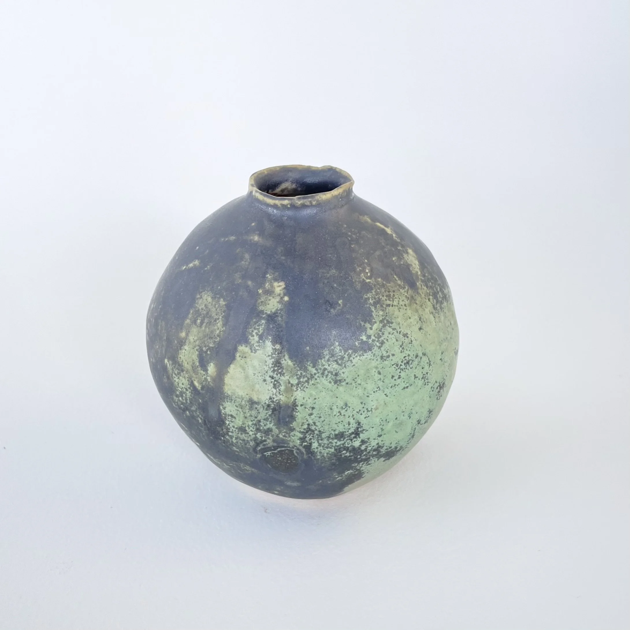 A round, black ceramic vase with a narrow opening at the top, featuring a surface with patches of green.