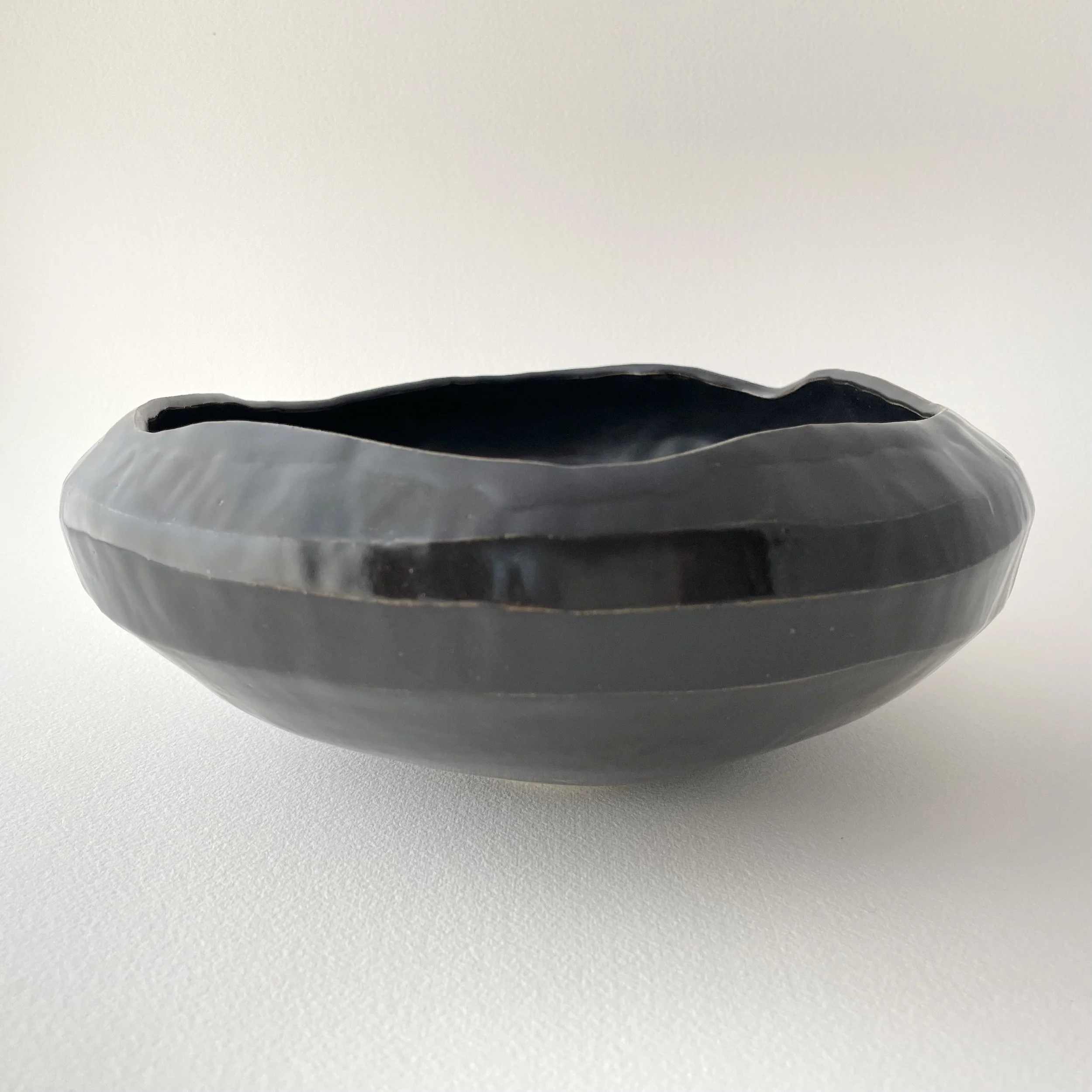 A black, shallow, round ceramic bowl with a matte finish and a slightly irregular rim, placed on a white surface.