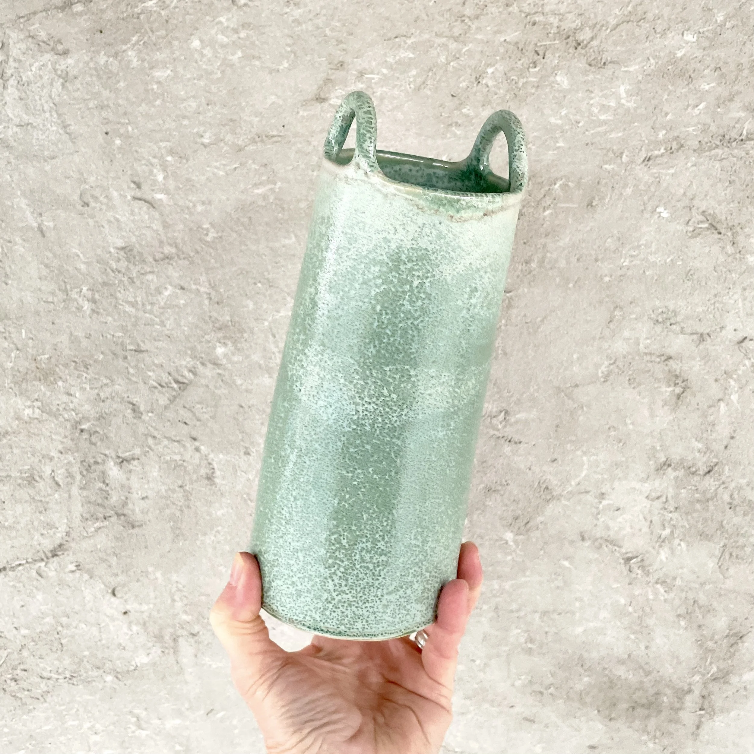 A person holds a tall, green, speckled ceramic vase with two loop handles against a textured beige wall.