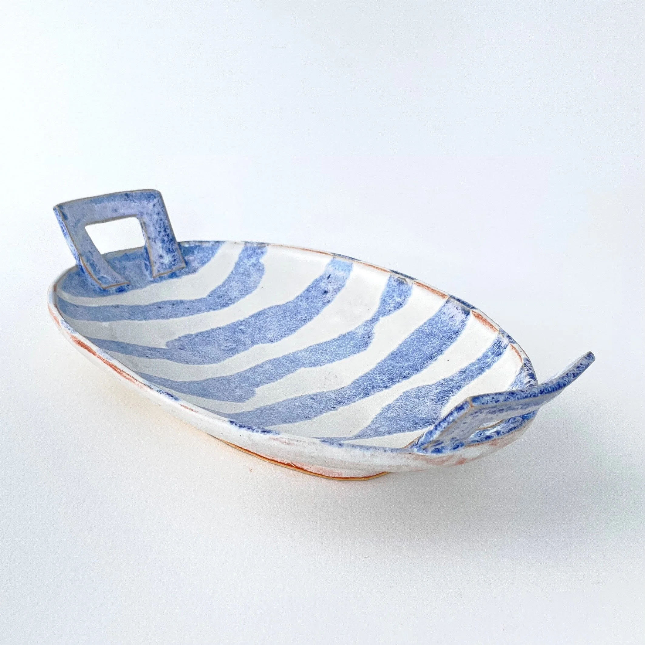 Small blue and white striped ceramic boat-shaped dish with two square handles on opposite sides, placed on a white surface.