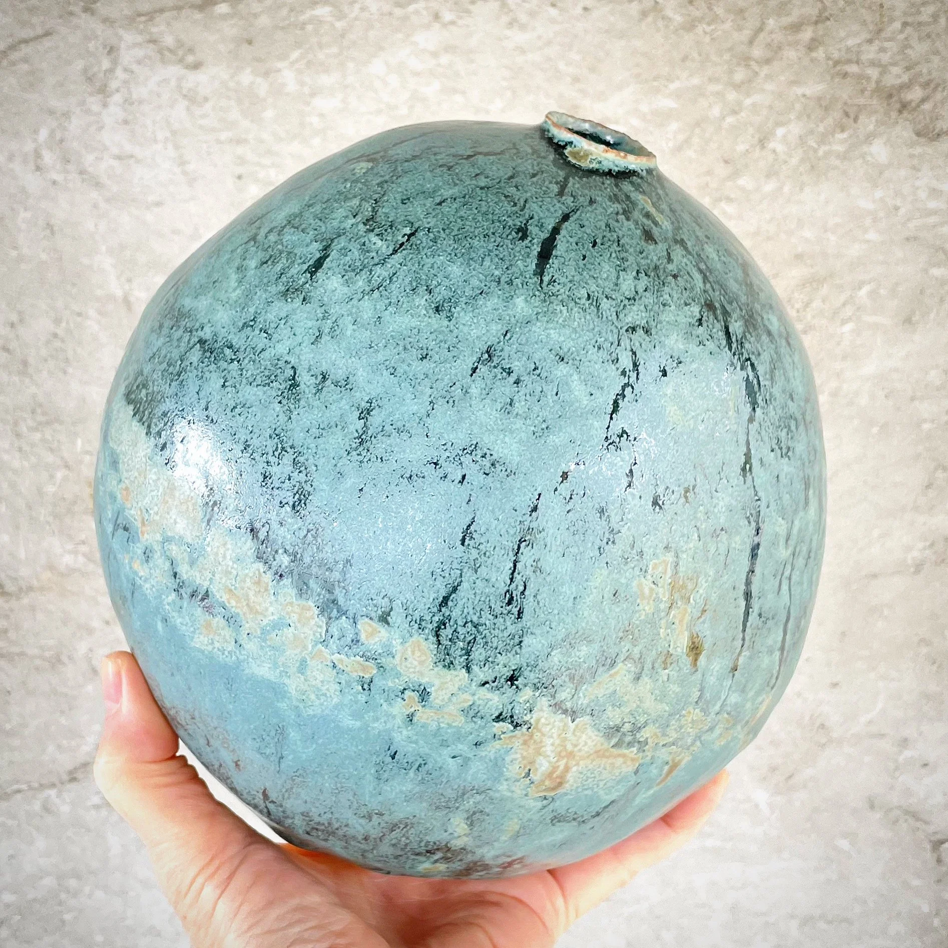 Person holding a round ceramic vase with a textured, mottled blue-green glaze and black and brown streaks, against a light beige wall.