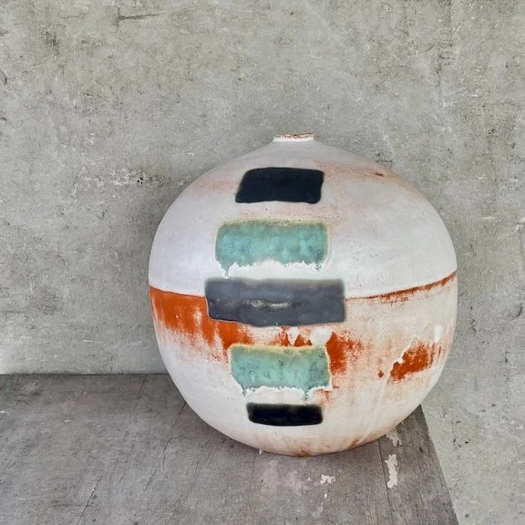 Large round ceramic orb with abstract colorful rectangular patterns, placed against a gray concrete wall.