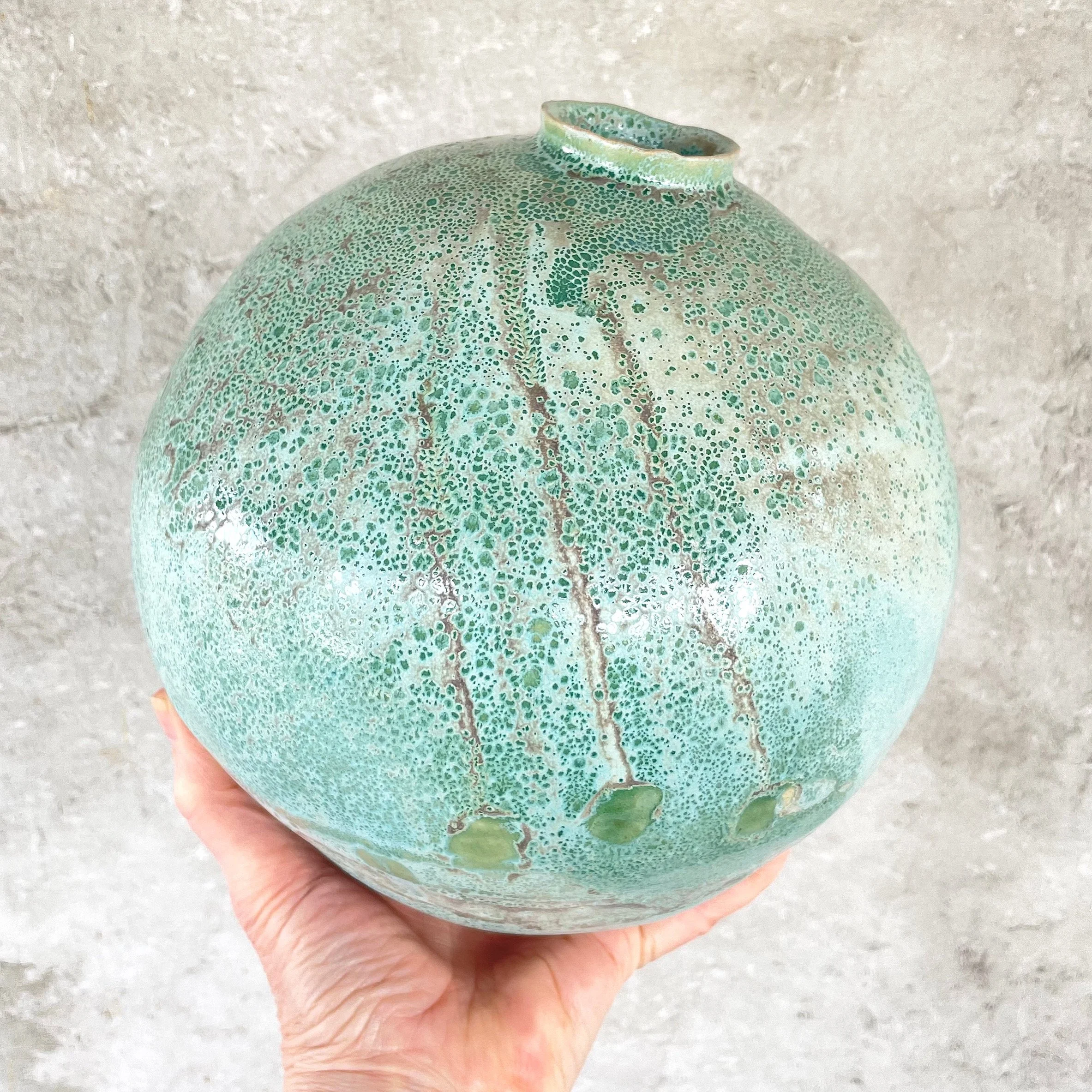 Hand holding a round, green ceramic orb vase against a light, textured background.