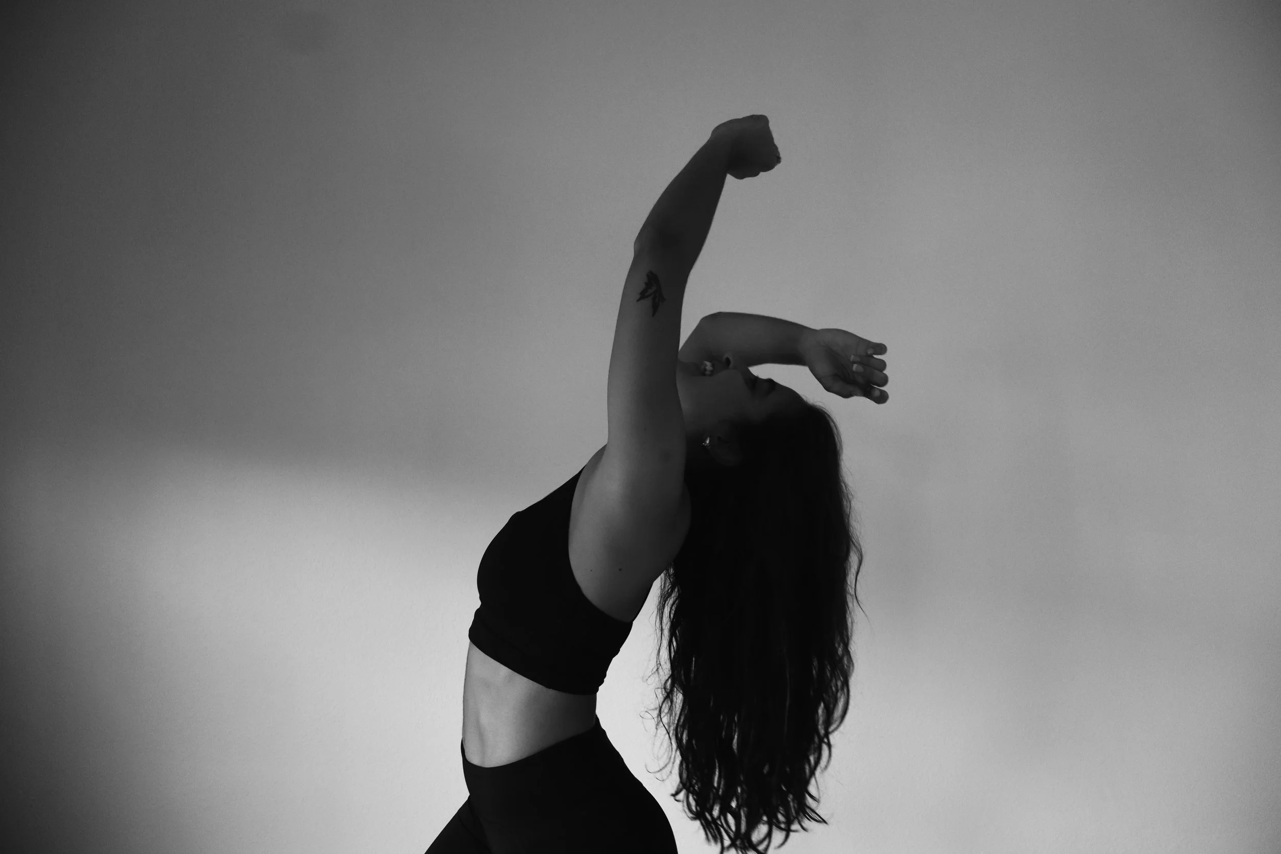 A woman with long hair in a black sports bra and leggings performing a dance or yoga pose against a plain background.
