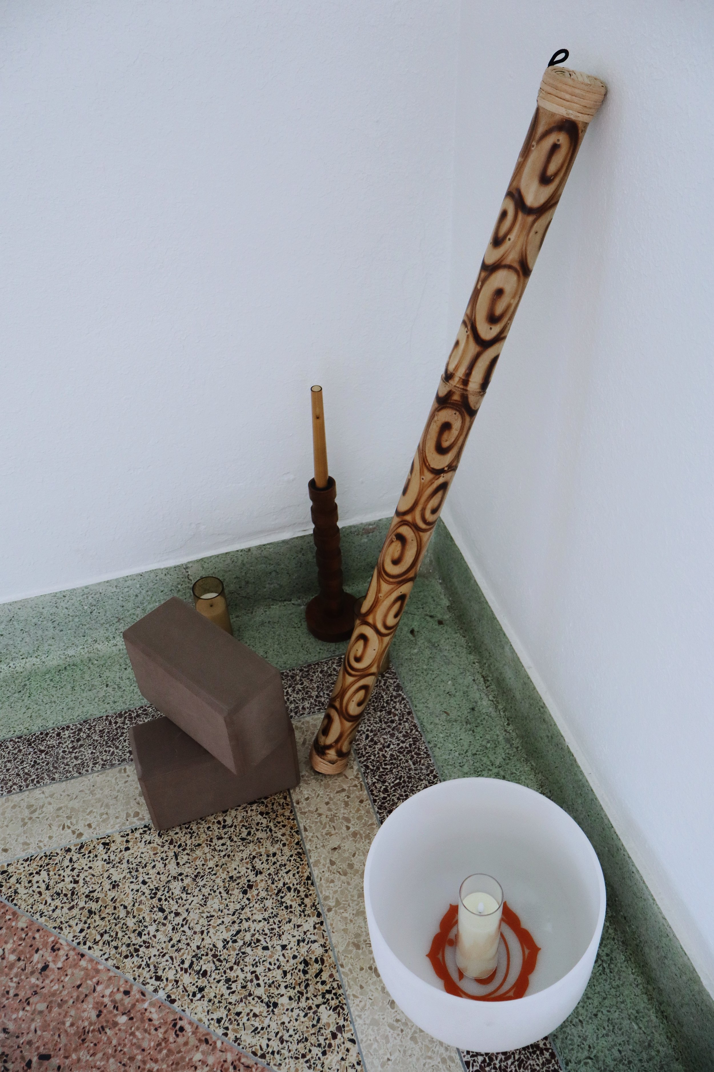 A small altar with a burning candle in a white holder with orange design, a brown block, a tall wooden pole with decorative swirl pattern, a wooden stick, and a small cylindrical object, all set against white walls and tile floor.