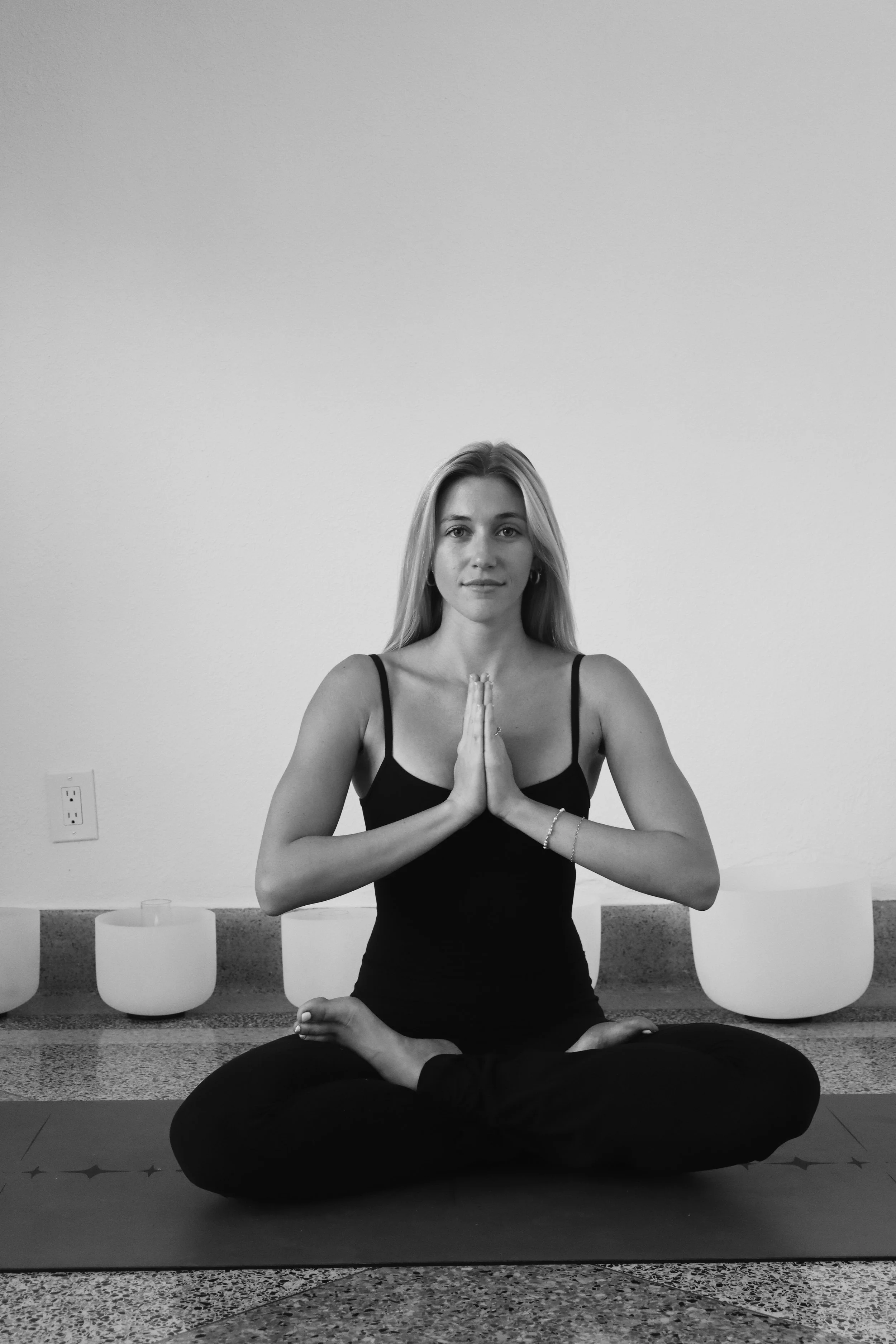 A woman practicing yoga in a cross-legged seated position with hands pressed together in prayer pose in front of her chest, sitting on a yoga mat, with singing bowls in the background.