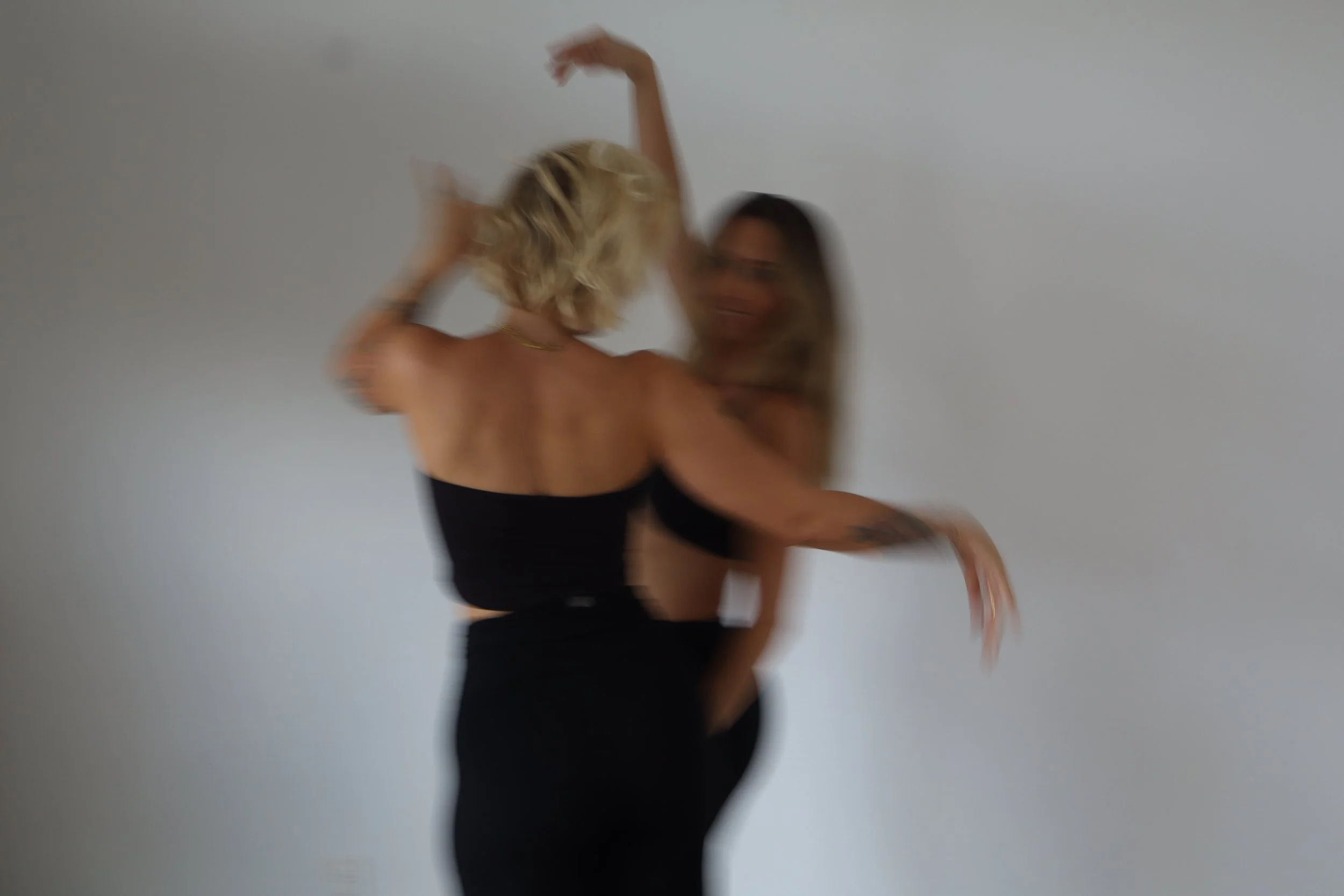 Two women dancing together against a plain white background, both smiling and wearing black outfits.