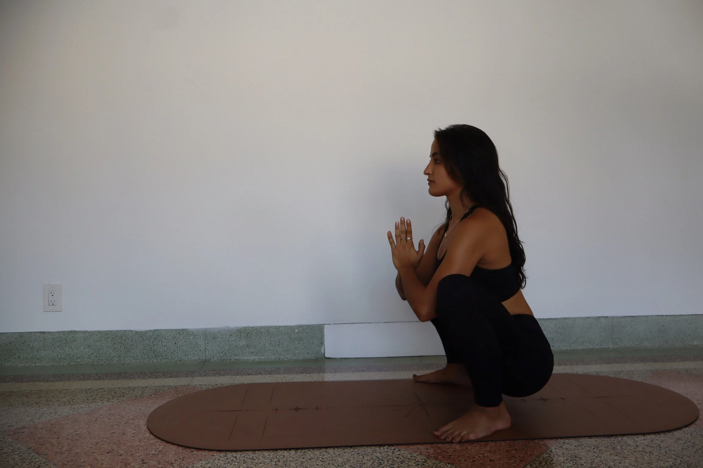 A woman in black workout clothes performing a yoga pose on a brown mat against a plain light-colored wall.