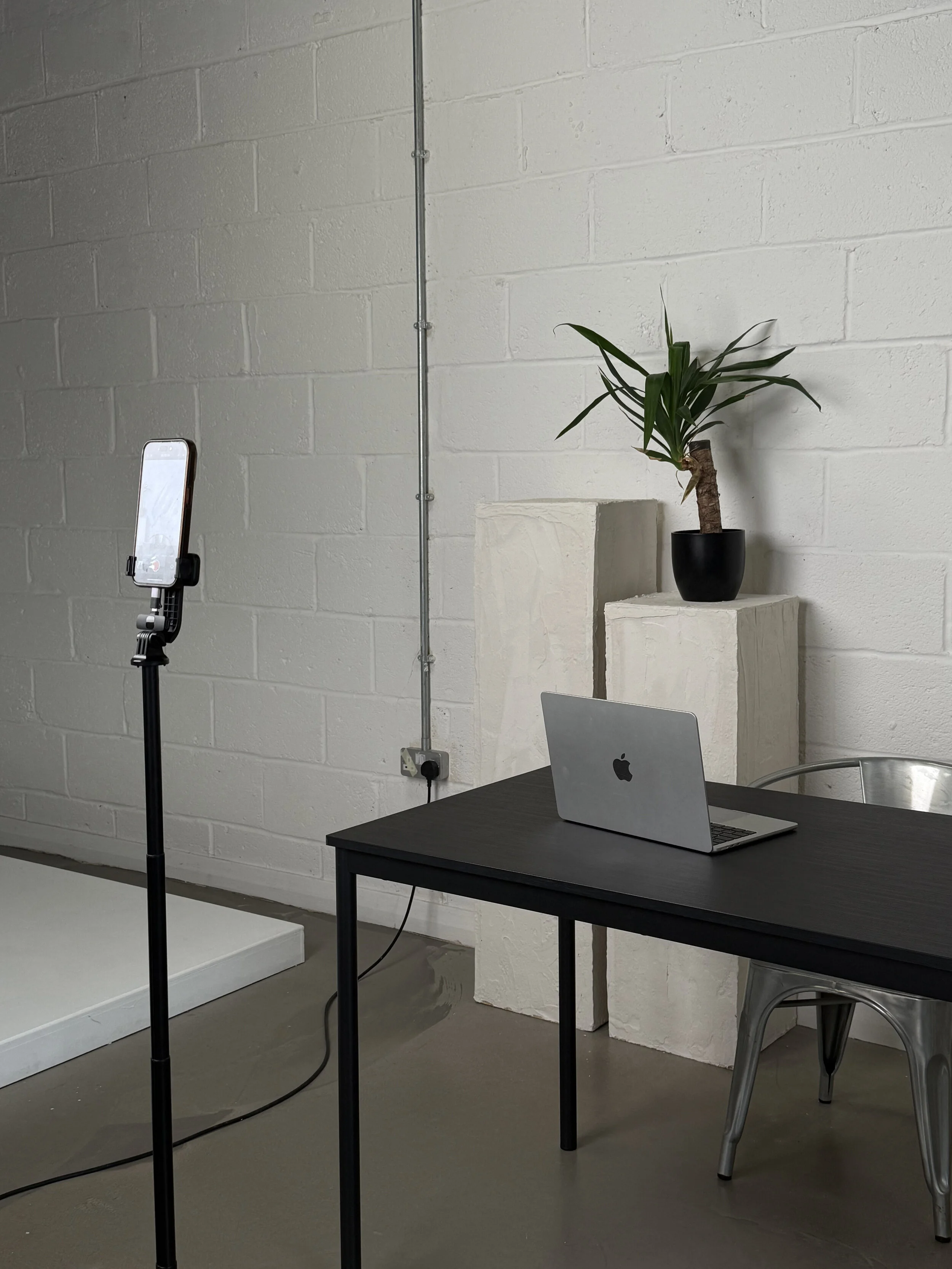 A photo of a black table with an open silver MacBook on it, a silver chair to the right, and a green potted plant on white pedestals against a white brick wall, with a smartphone on a tripod stand capturing the scene.