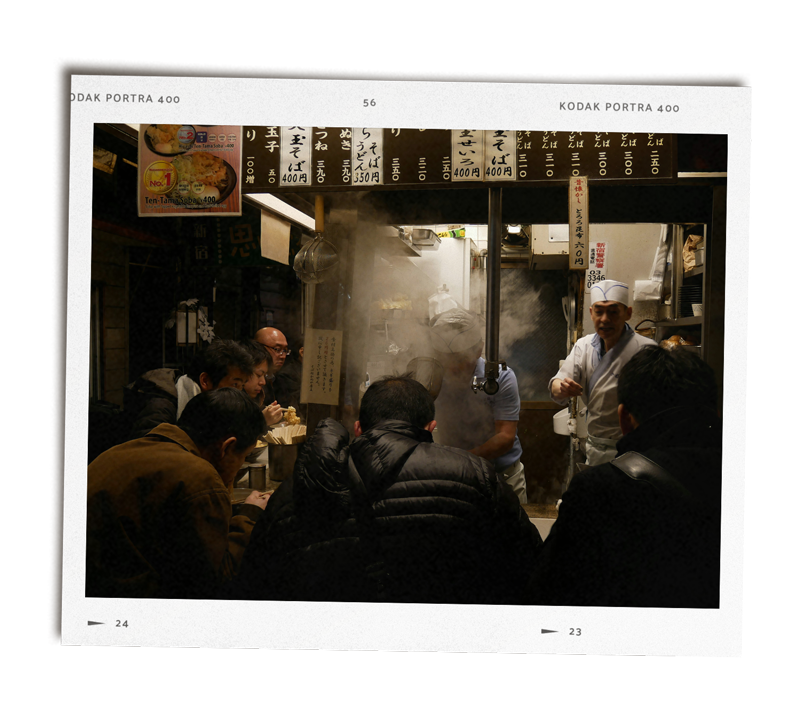 A busy Japanese ramen restaurant with customers seated closely together at the counter, chefs preparing food behind the steam and smoke, with Japanese on the menu and signs visible.