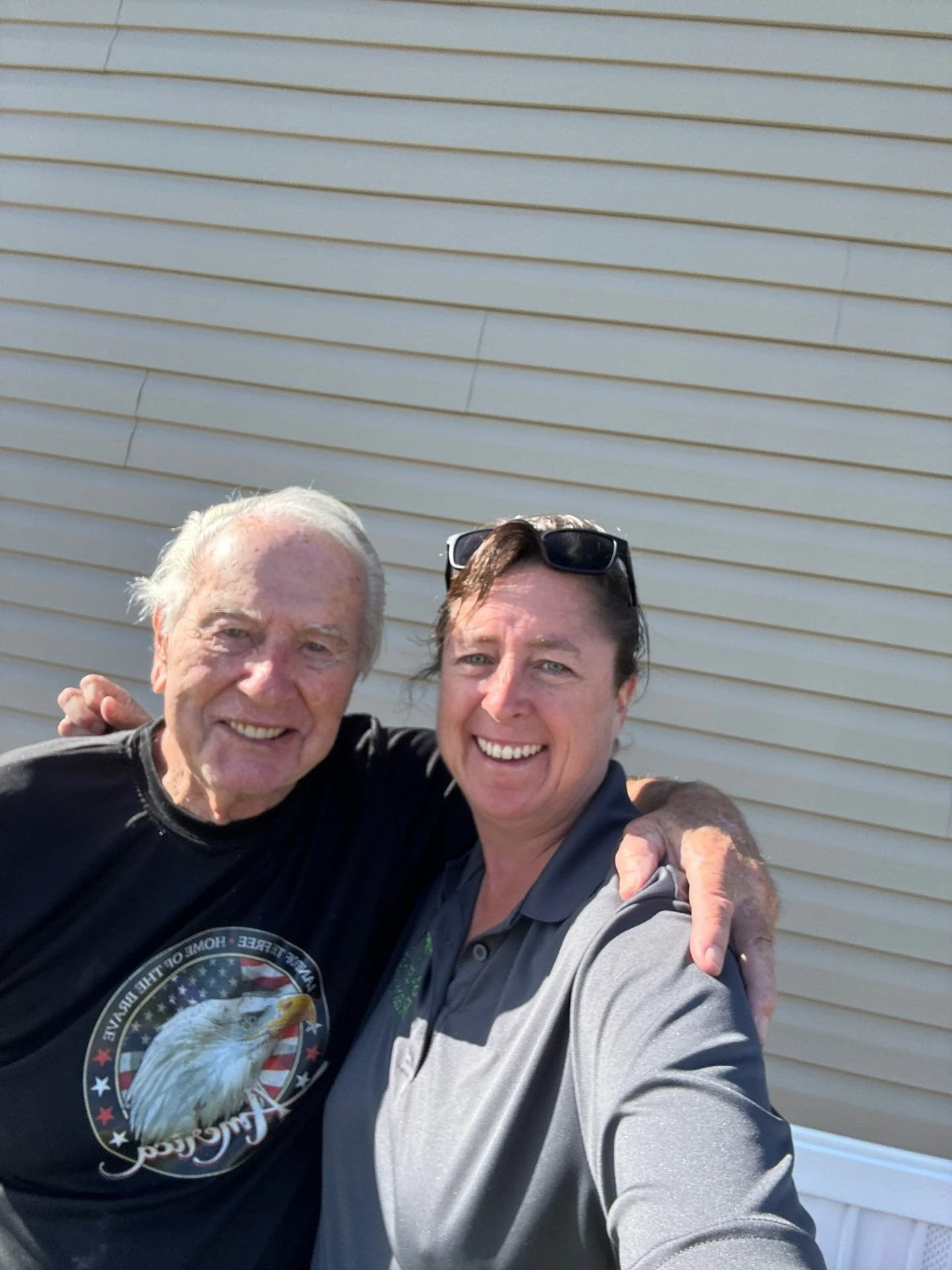 Two smiling men, one elderly with white hair and one middle-aged with dark hair, taking a selfie outside against a beige siding wall. The elderly man is wearing a black T-shirt with an eagle and American flag design, while the other man wears a gray
