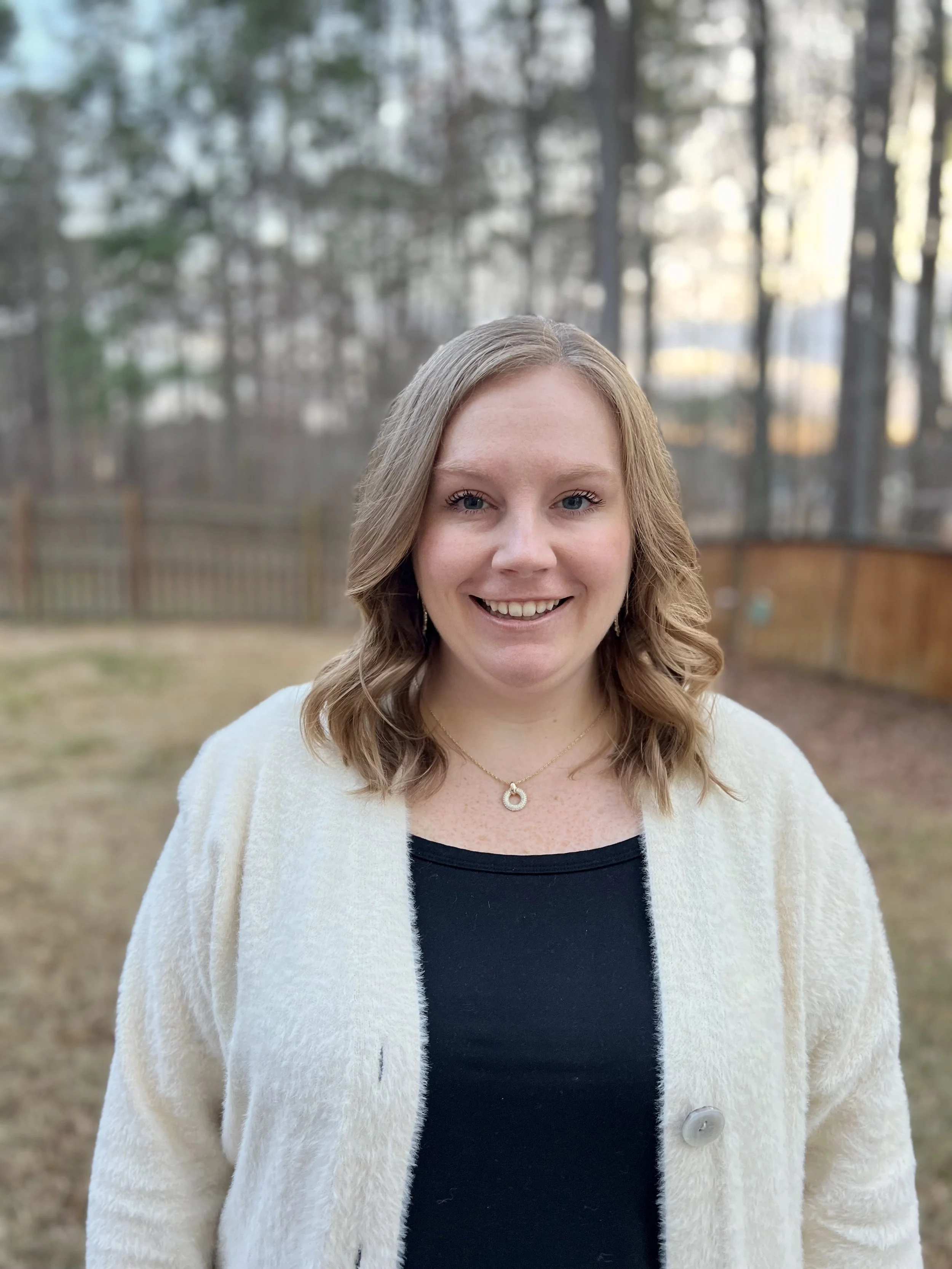 A woman with shoulder-length wavy blonde hair, smiling, standing outdoors in a backyard with a wooden fence and trees in the background. She is wearing a cream-colored cardigan over a black top and a gold necklace with a circular pendant.