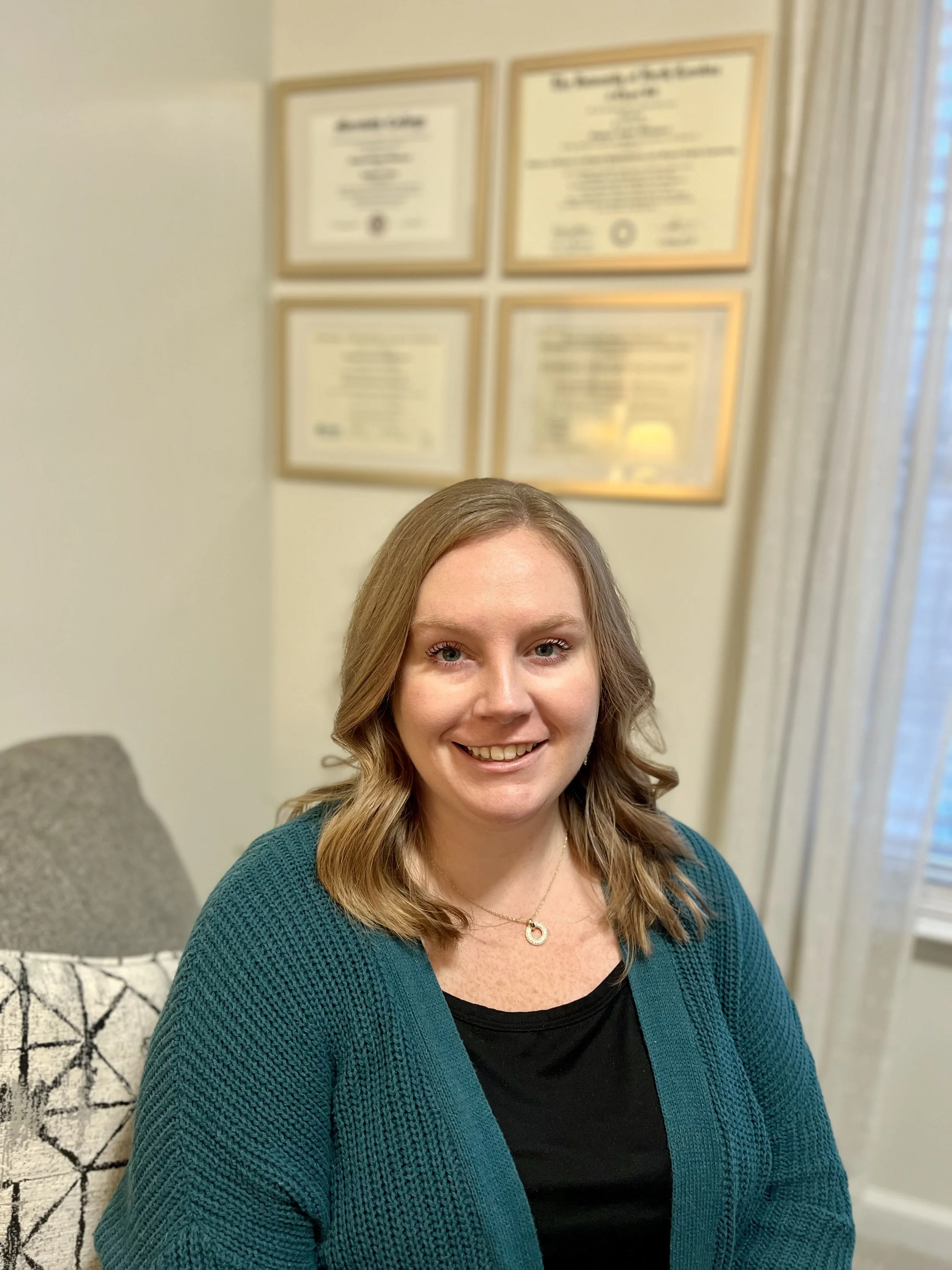 A woman with shoulder-length light brown hair, smiling, wearing a black top and teal cardigan. She is seated indoors with framed certificates on the wall behind her and a window with sheer curtains to the side.