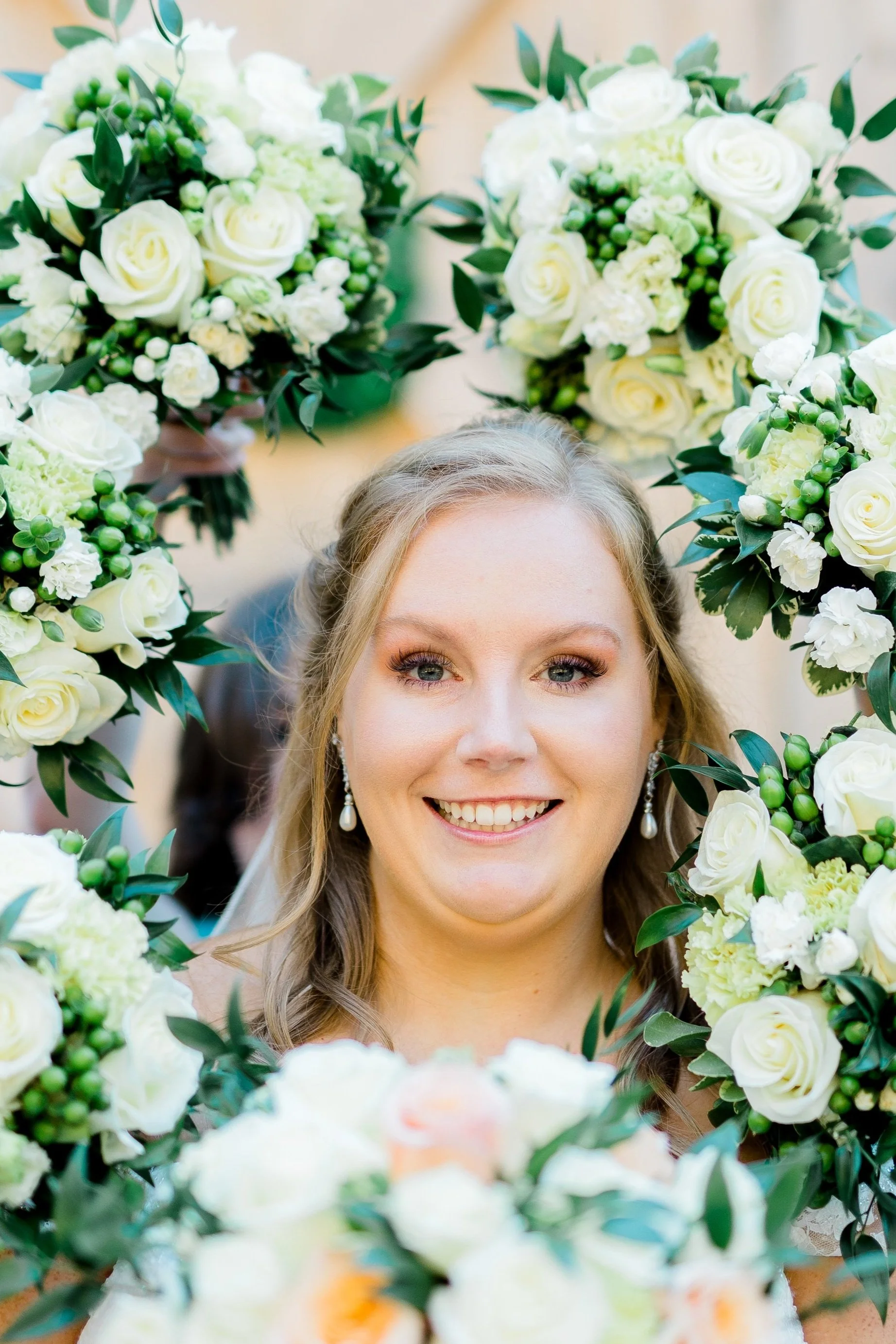 A smiling bride with earrings surrounded by white and green floral arrangements.