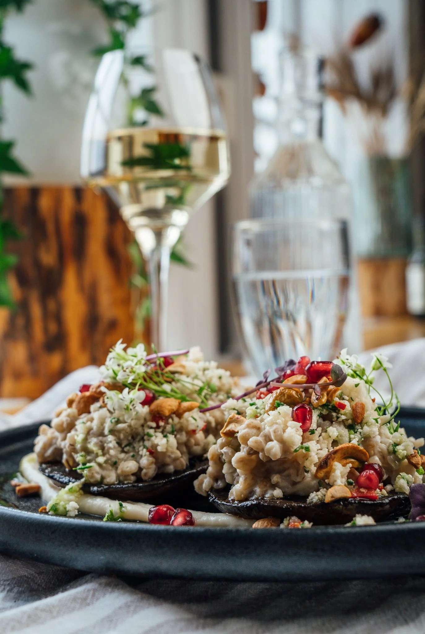 Two stuffed mushroom caps topped with chopped nuts, red pomegranate seeds, microgreens, and cheese on a dark plate with a glass of white wine and water in the background.