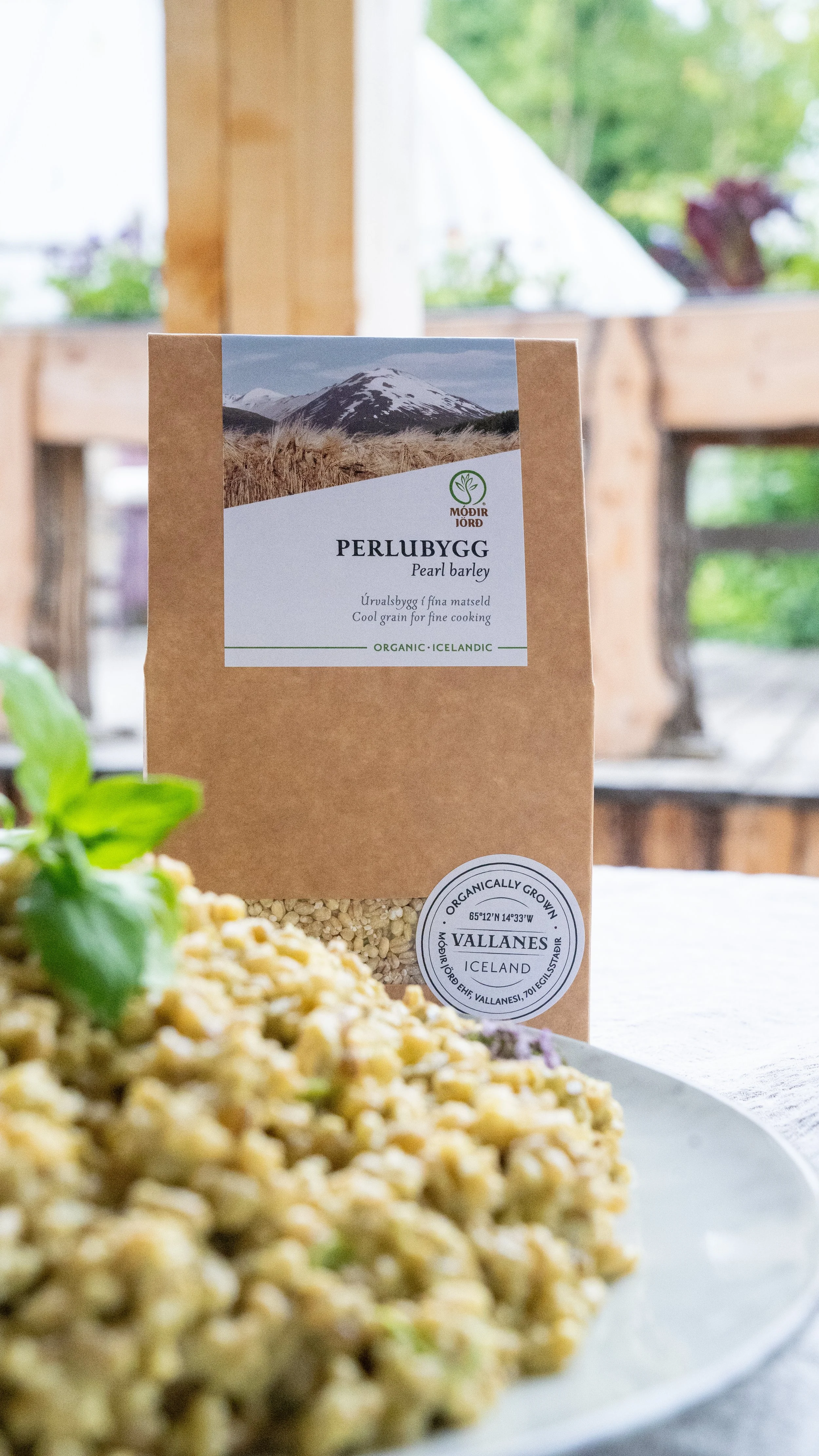 Close-up of a package of pearl barley labeled 'Perlubygg' with an Icelandic mountain landscape on the label, placed on a table near a plate of cooked barley dish.