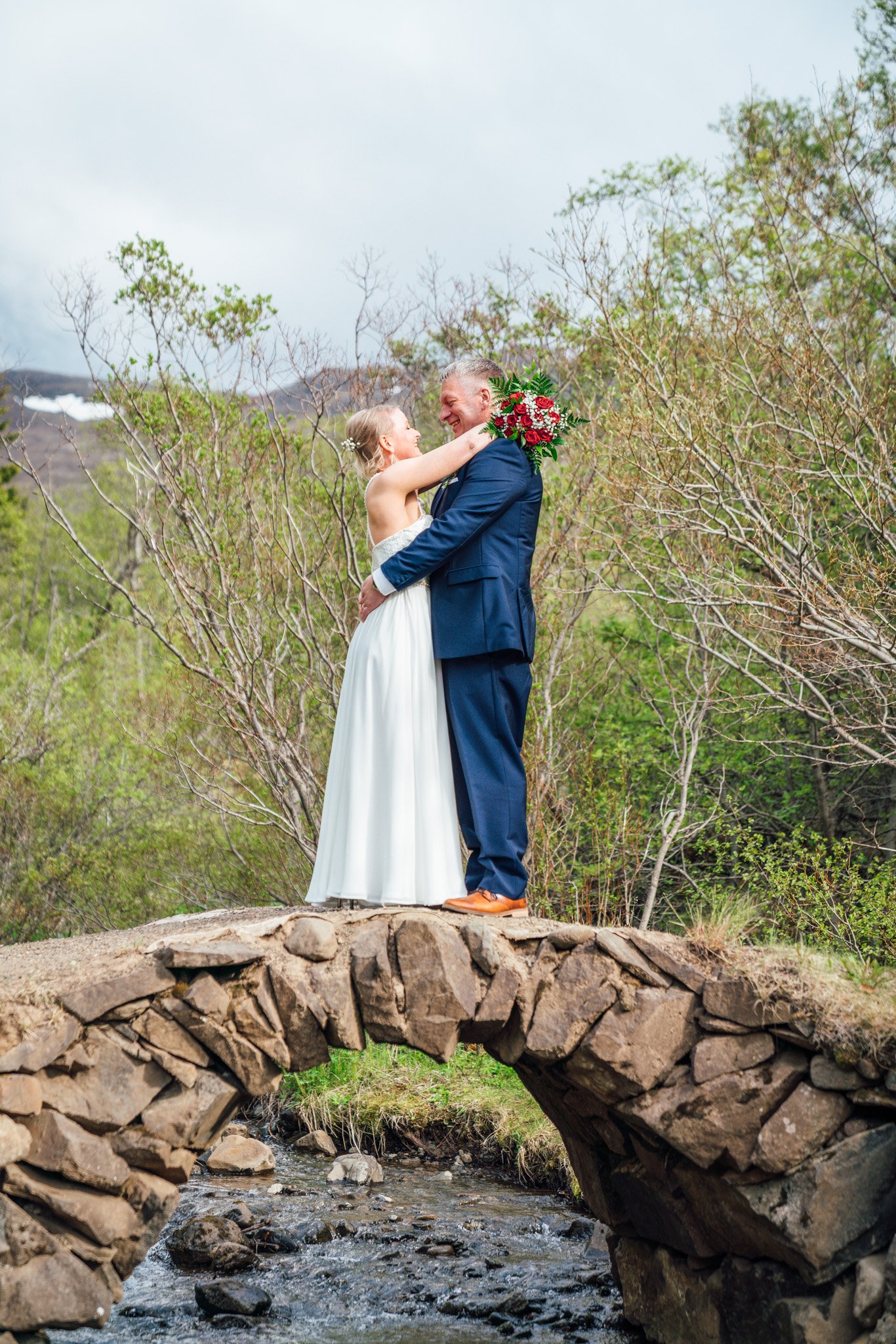 A bride and groom standing on a small stone bridge over a stream, embracing and smiling, outdoors with trees and mountains in the background.