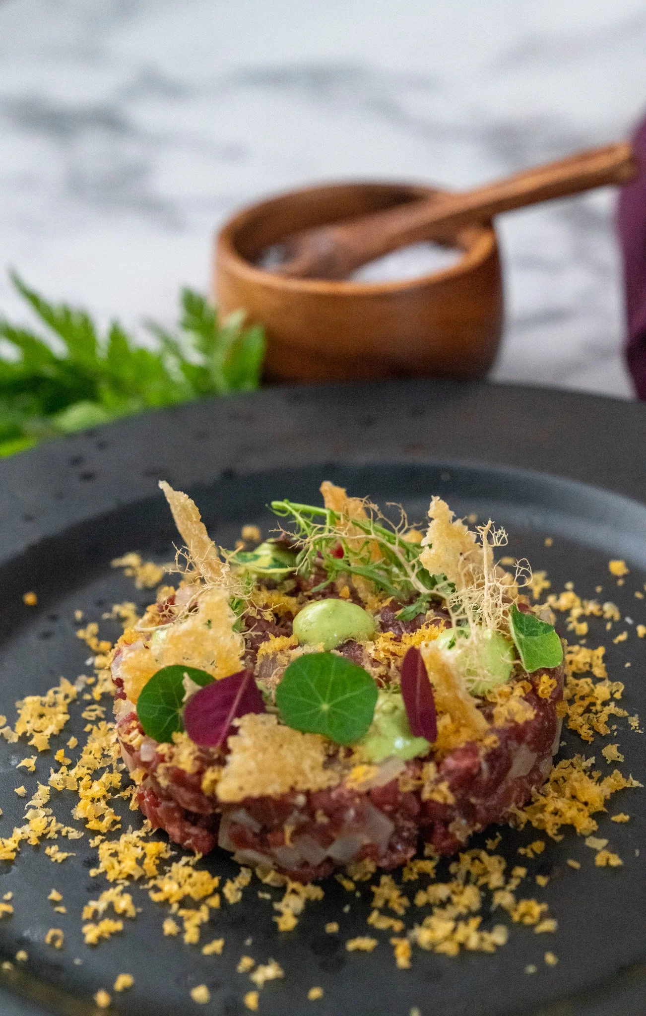 A plated gourmet dish featuring diced raw beef, garnished with microgreens, crispy elements, and green sauces, with a wooden bowl of salt and a spoon in the background.