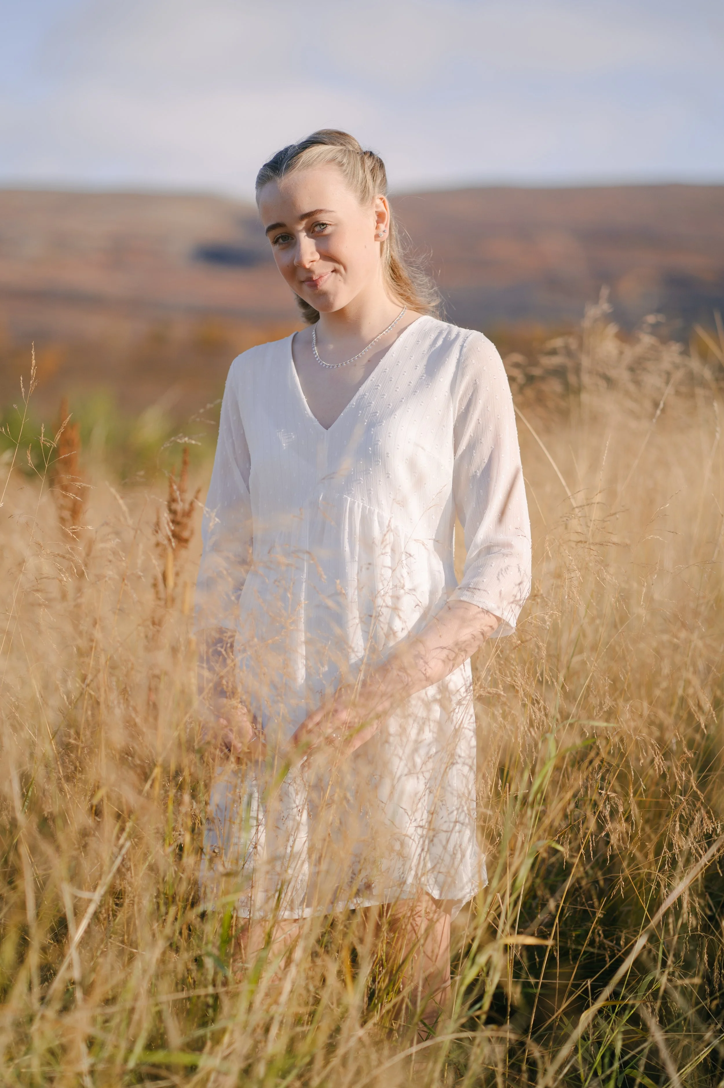 A young woman in a white dress standing in a field of tall, golden grass with hills in the background on a sunny day.