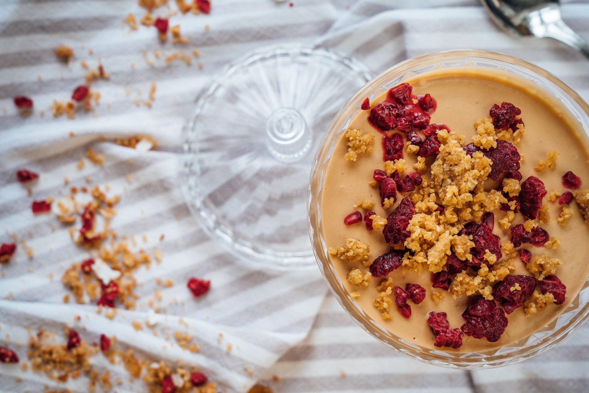Top view of a glass dessert dish filled with creamy pudding topped with dried cranberries and crushed graham crackers, placed on a textured fabric with scattered crumbs and pieces of dried cranberries.